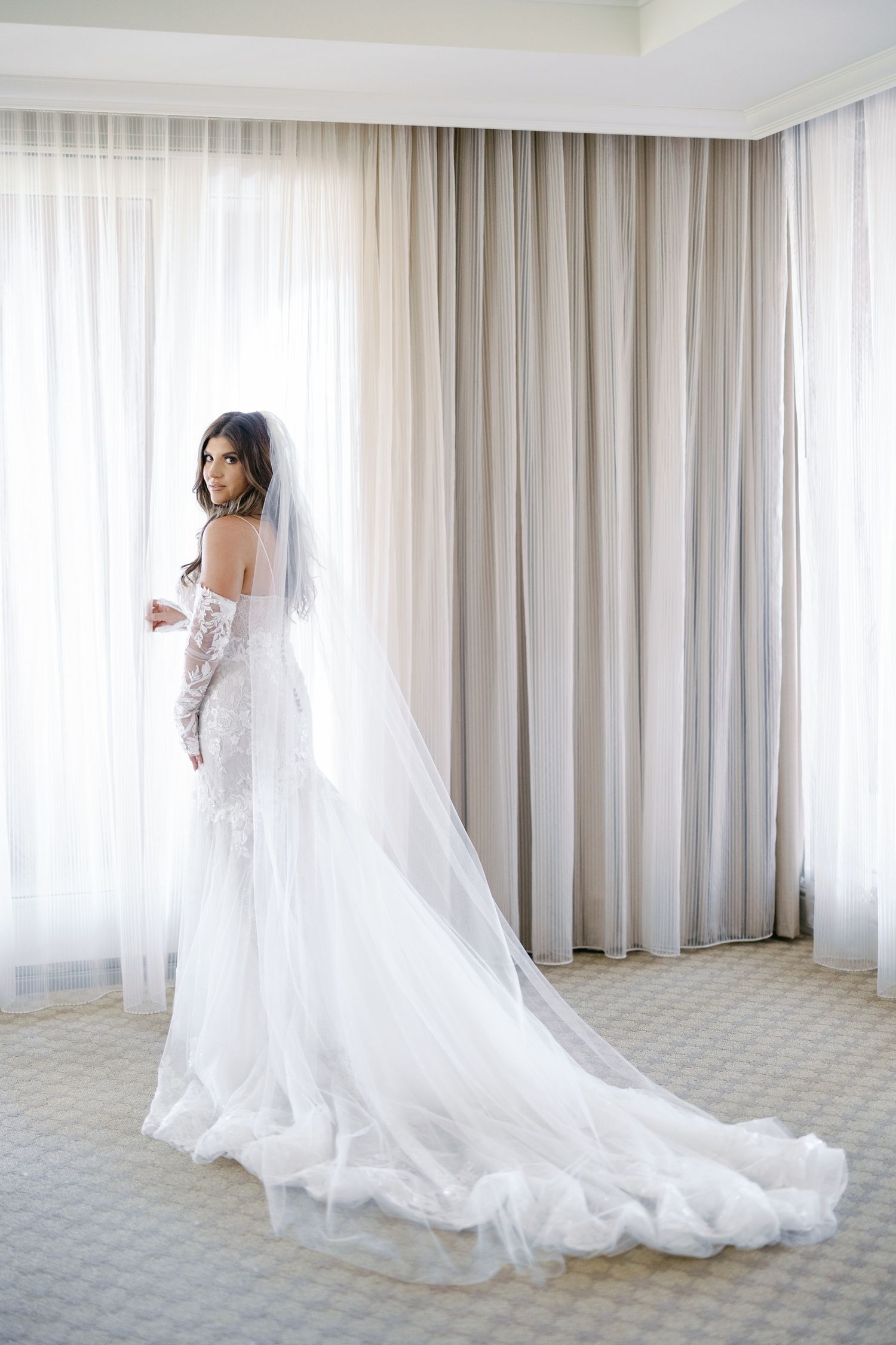 Bride in elegant lace mermaid gown with long cathedral veil stands by draped curtains in bridal suite.