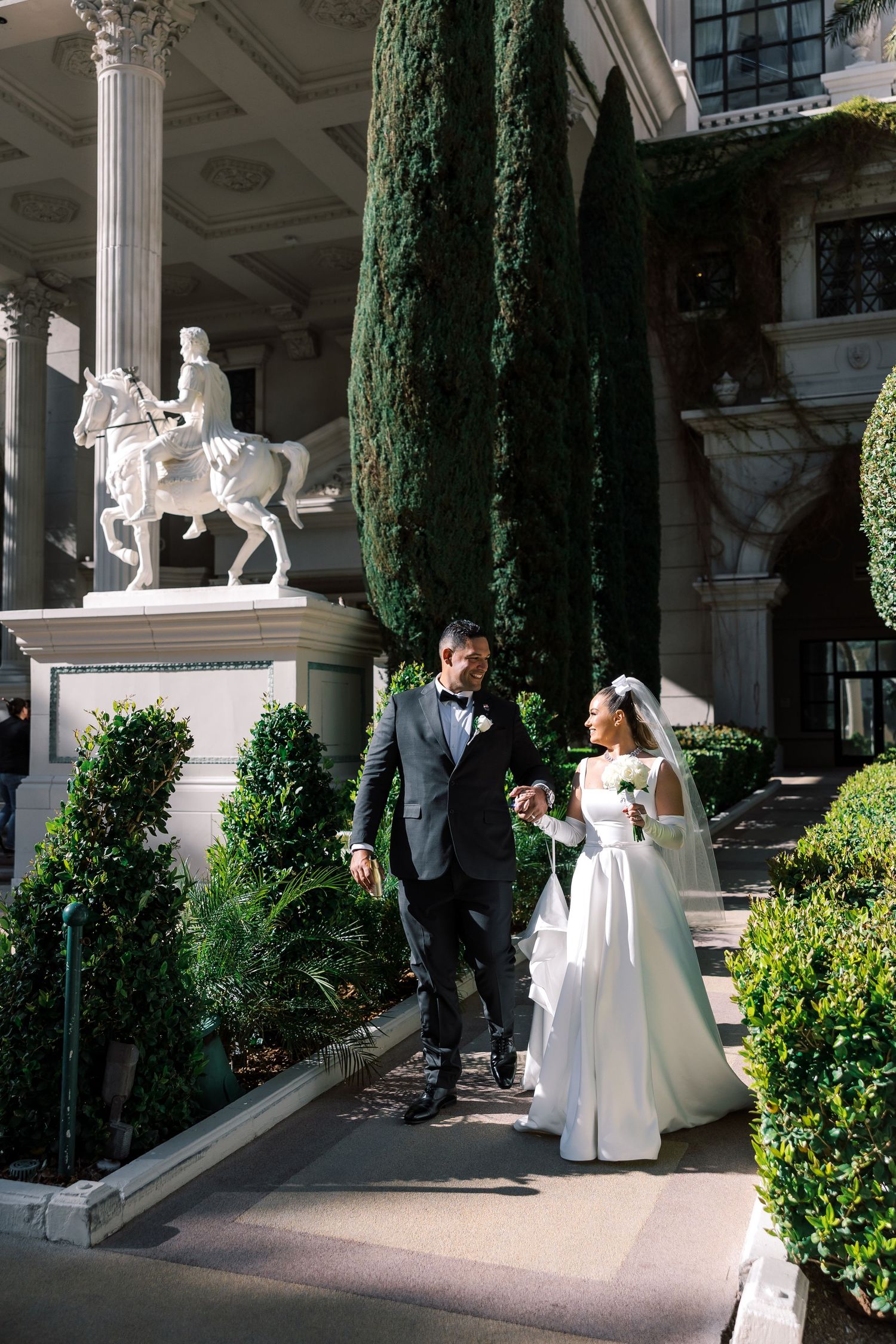 Bride and groom walk hand-in-hand outside a grand venue with tall cypress trees and a white equestrian statue.
