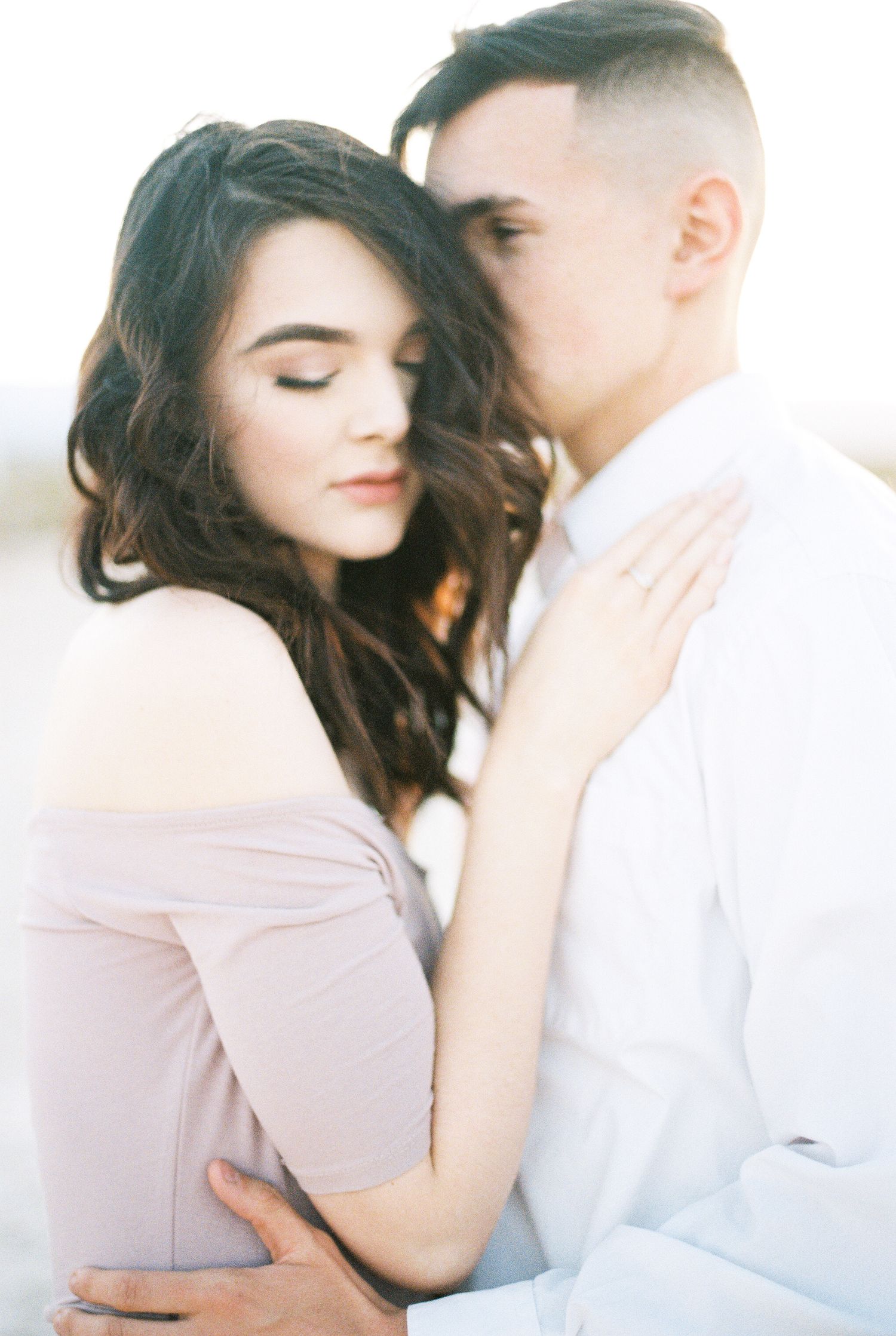 A couple shares an intimate moment, the man kissing the woman's temple as she closes her eyes in a soft embrace.