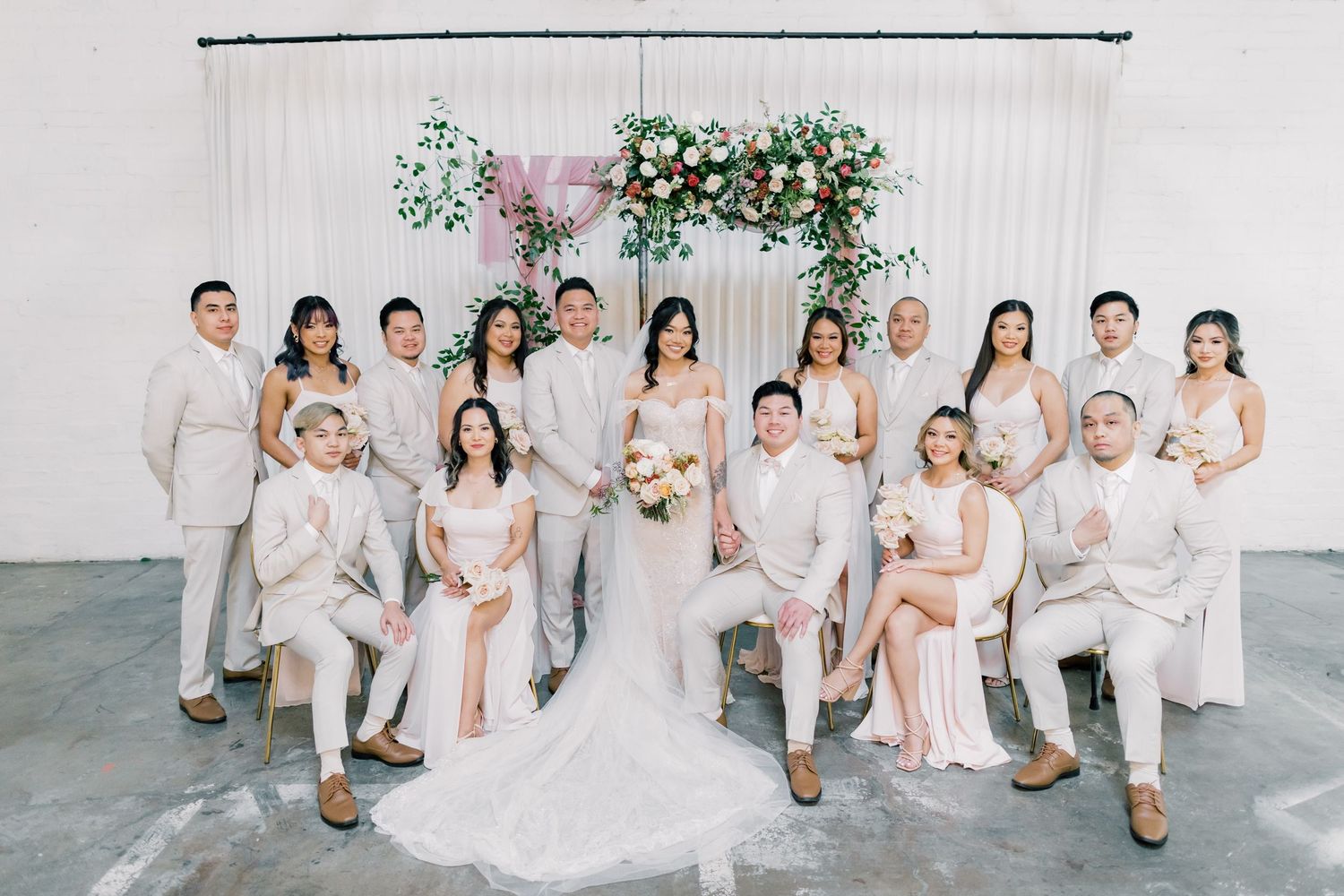 Wedding party in coordinated white and beige outfits poses together for formal photos against floral backdrop.
