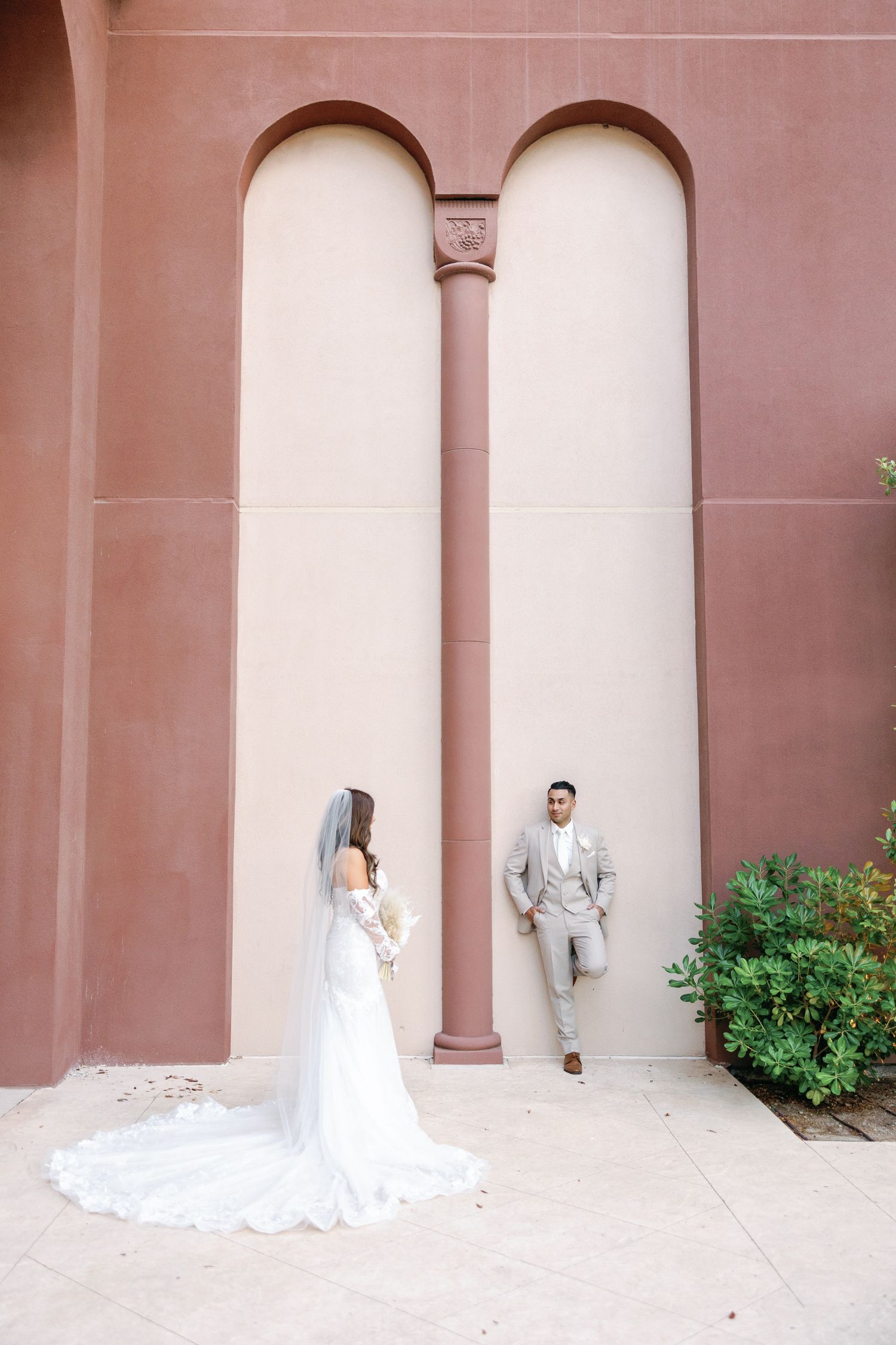 Bride in white gown and groom in gray suit pose near arched columns of a terracotta building on their wedding day.