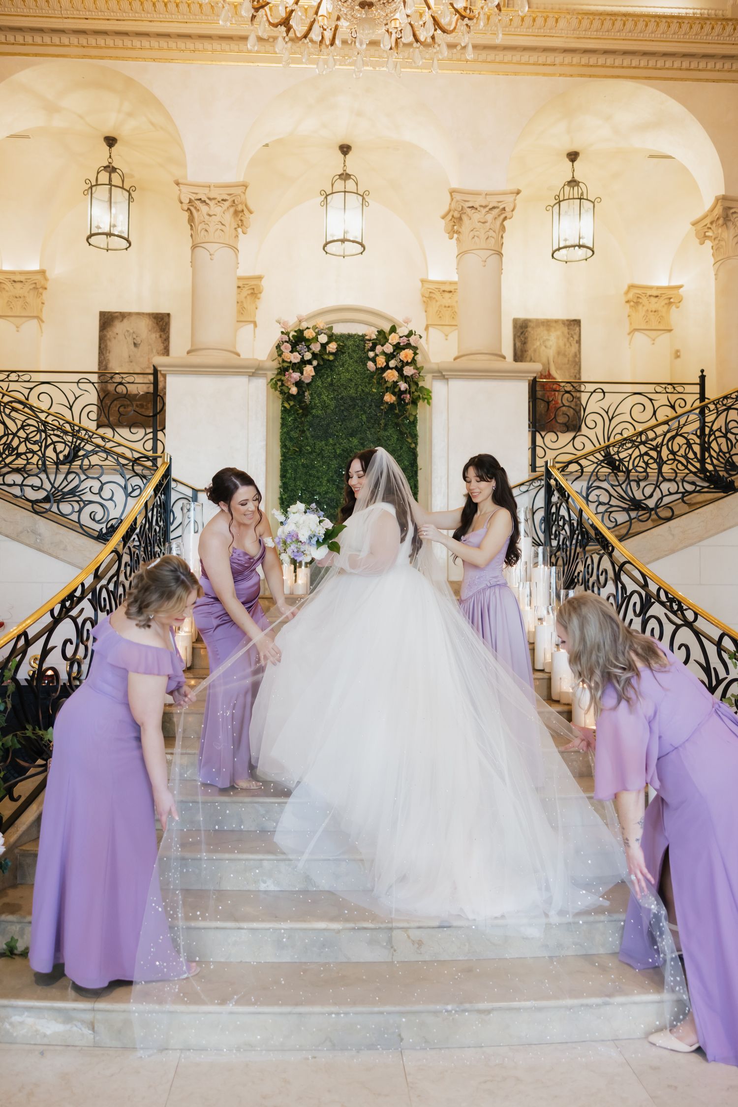Bride in white ballgown on grand staircase with bridesmaids in lavender dresses arranging her train.