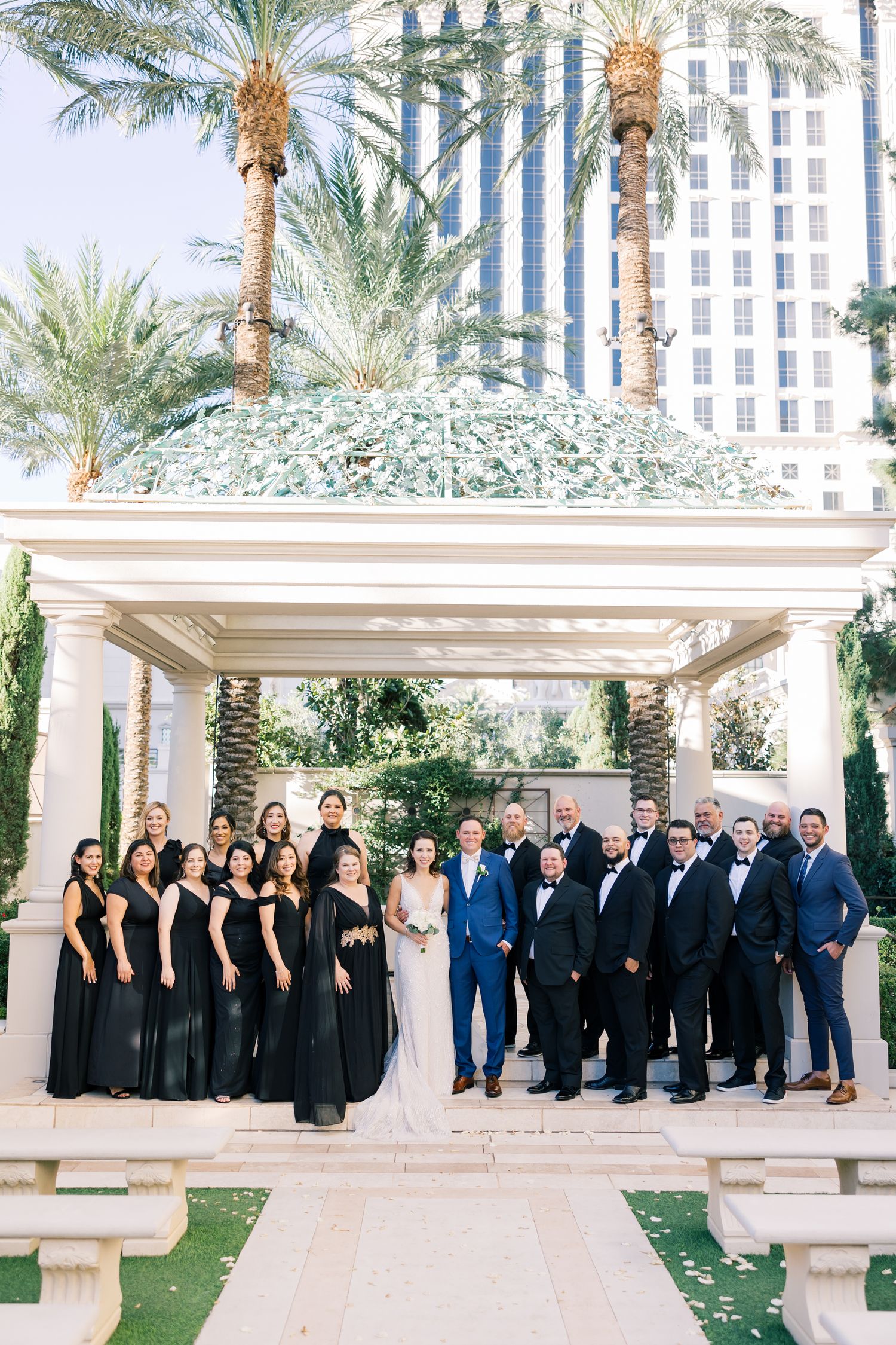 Wedding party in black dresses and tuxedos poses under pergola with palm trees and hotel in background.