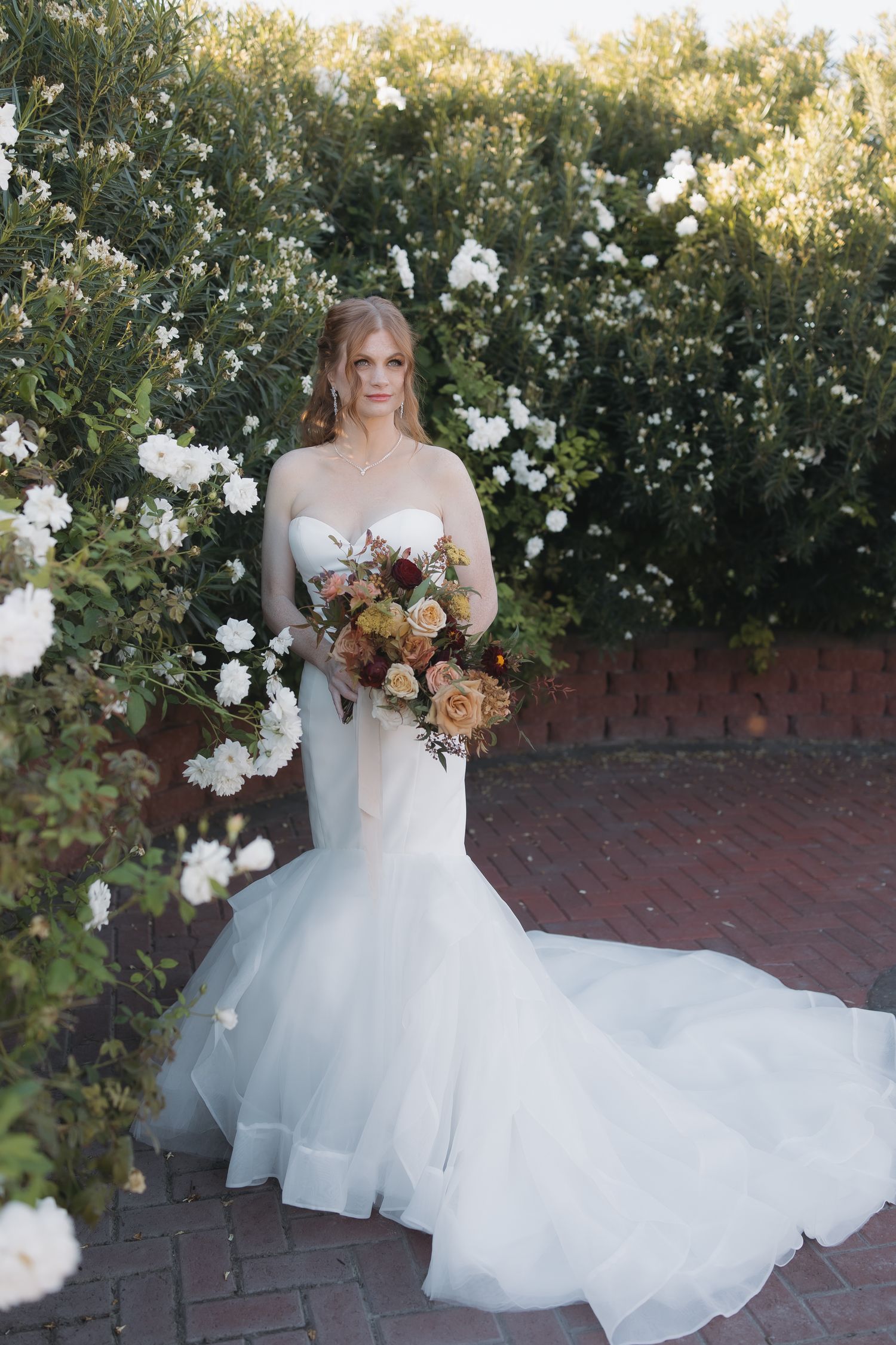 Bride in strapless mermaid wedding gown holding autumn bouquet, posing before lush white flowering bushes.