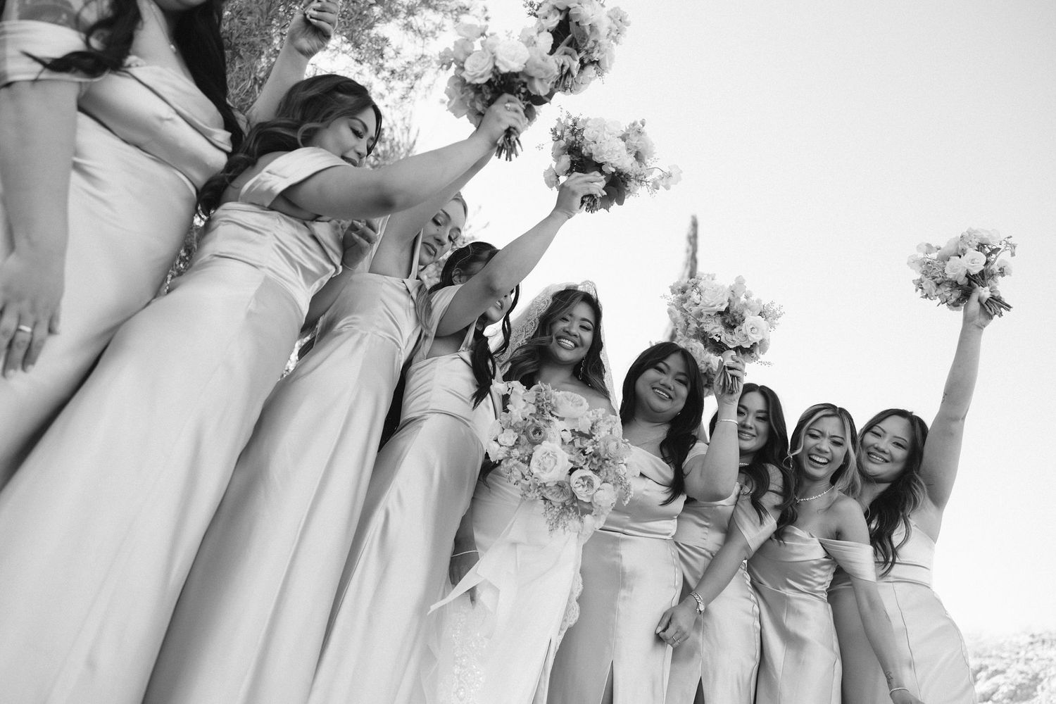 Joyful wedding party wearing white dresses poses together while holding bouquets of flowers in a candid celebration moment.