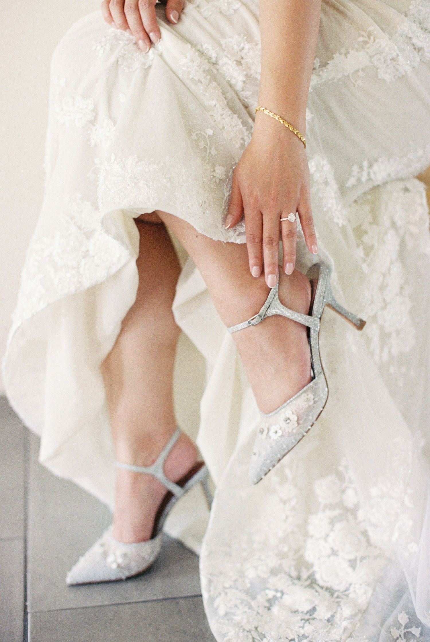 Bride in lace wedding dress adjusting silver embellished heels with ankle strap, wearing gold bracelet and diamond ring.