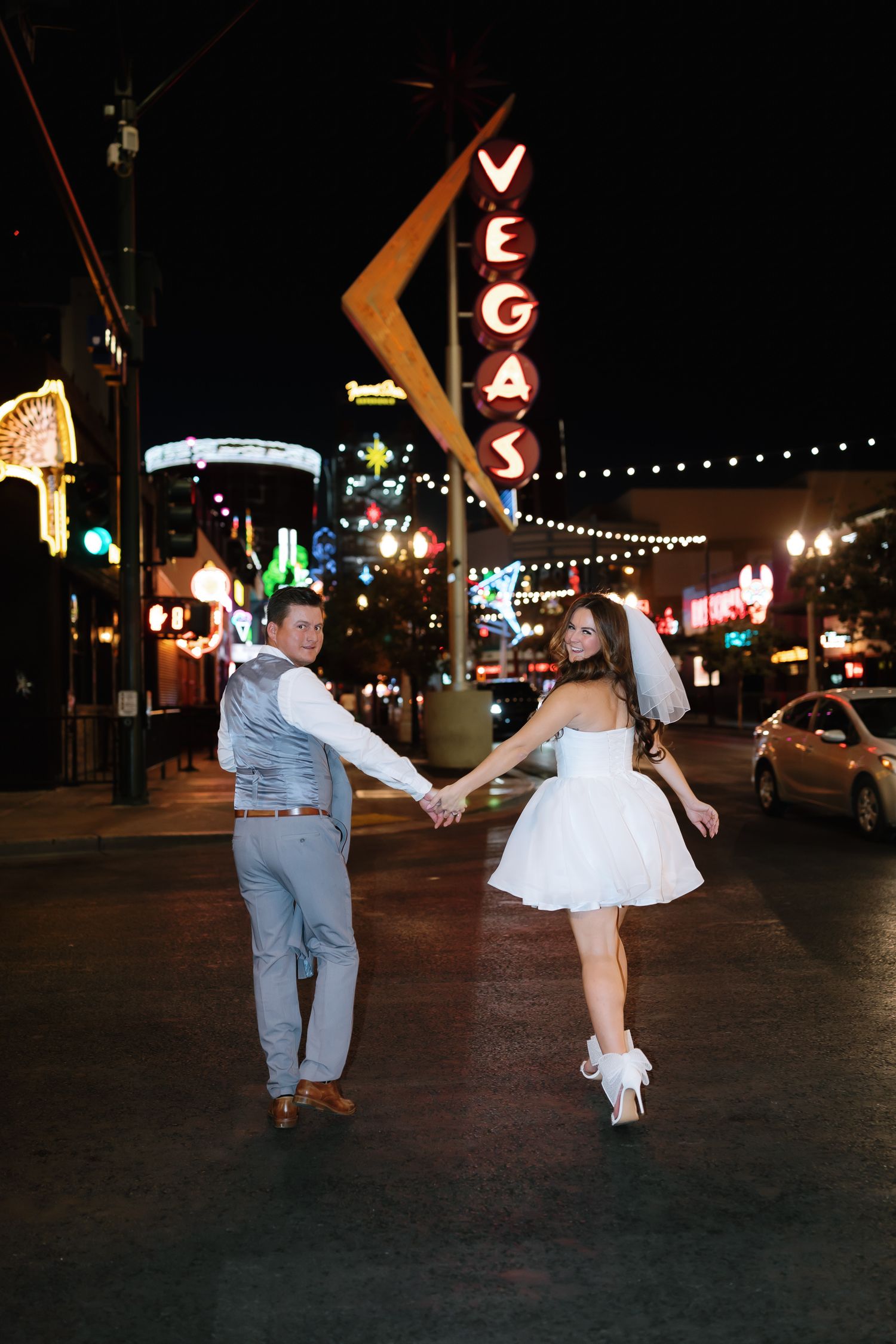 Newlywed couple holding hands on the Las Vegas Strip at night with glowing Vegas neon sign behind them.