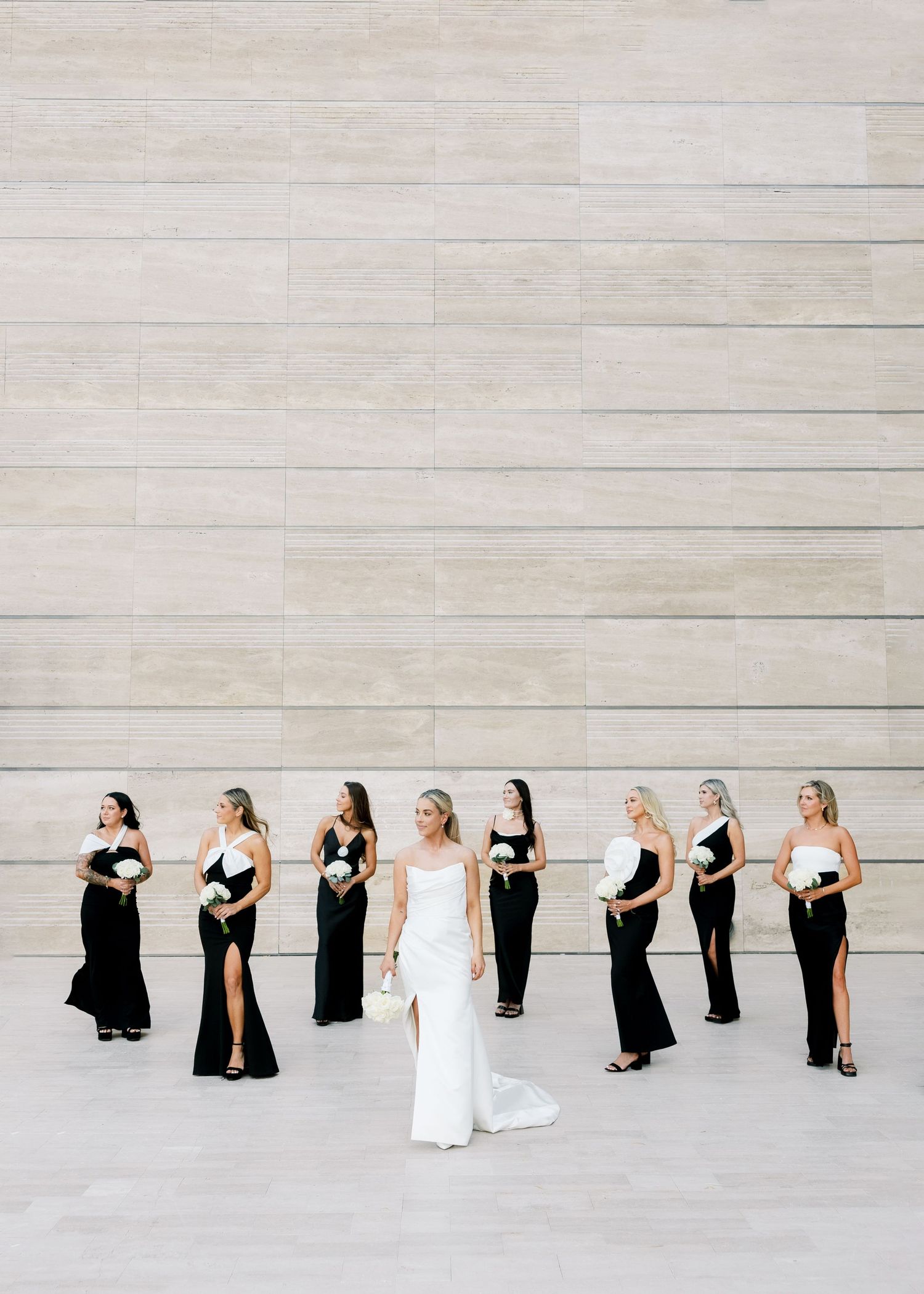 Bride in white gown poses with bridesmaids in black dresses against a limestone wall.
