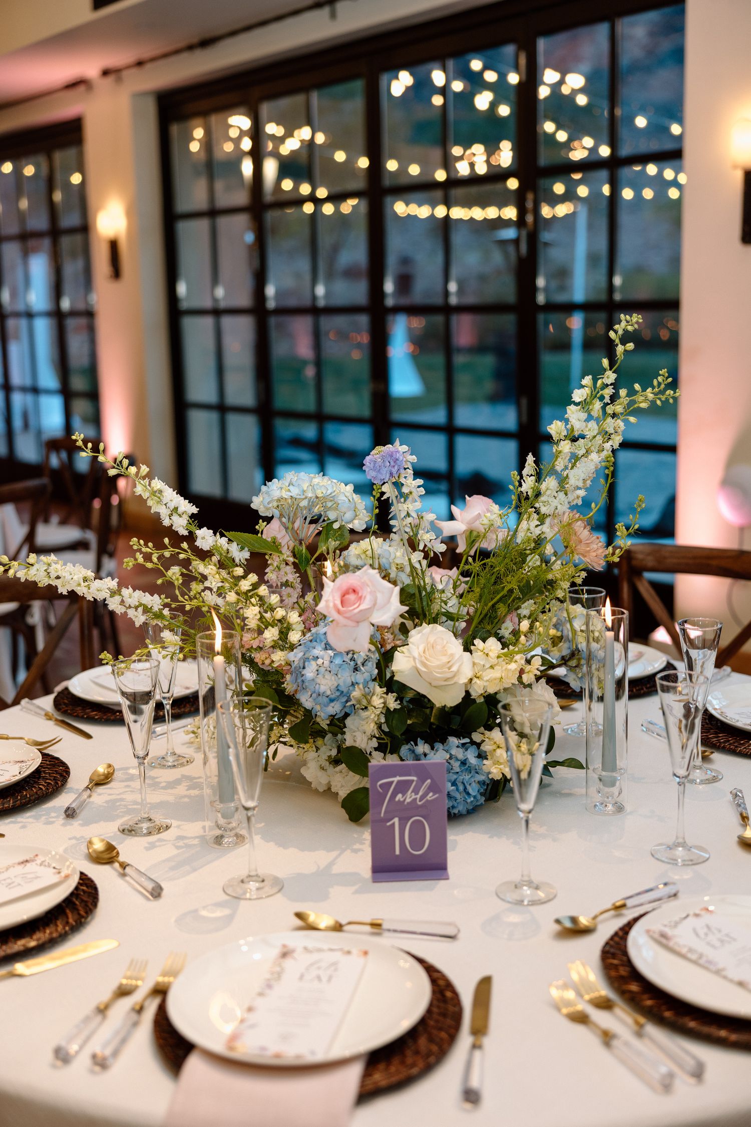 Elegant wedding reception table 10 with blue hydrangea floral centerpiece, crystal glasses, and wicker charger plates.