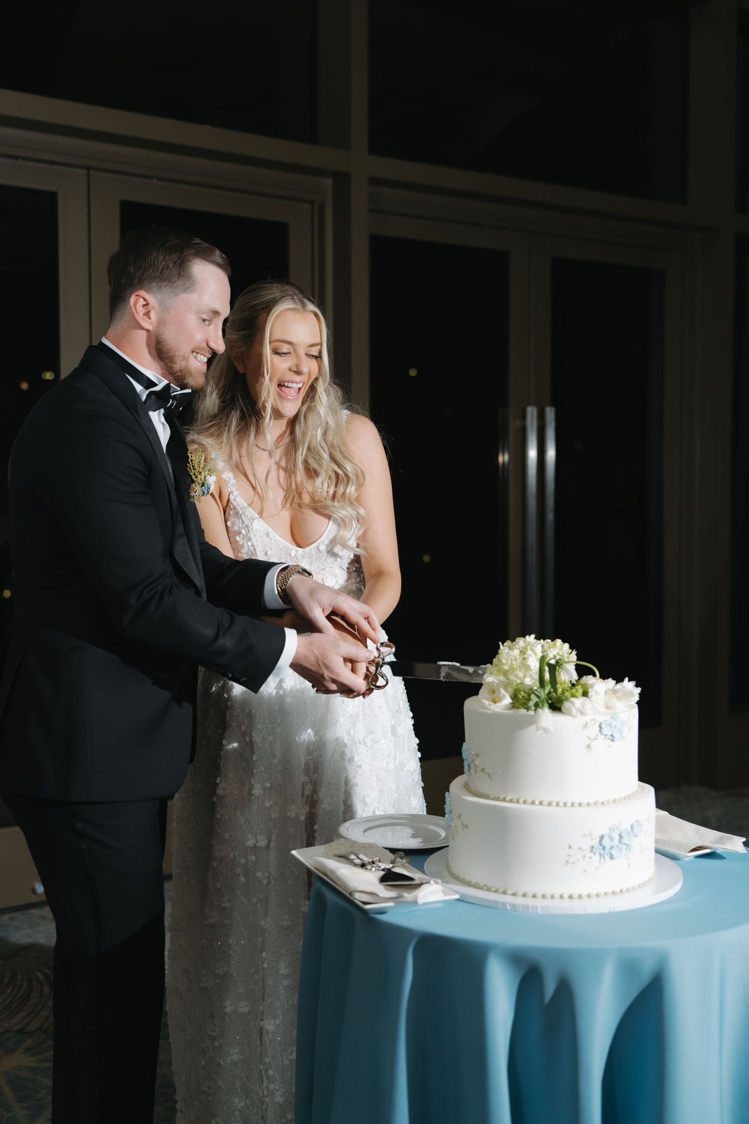 Smiling bride and groom cut their elegant two-tier white wedding cake with blue floral details together.