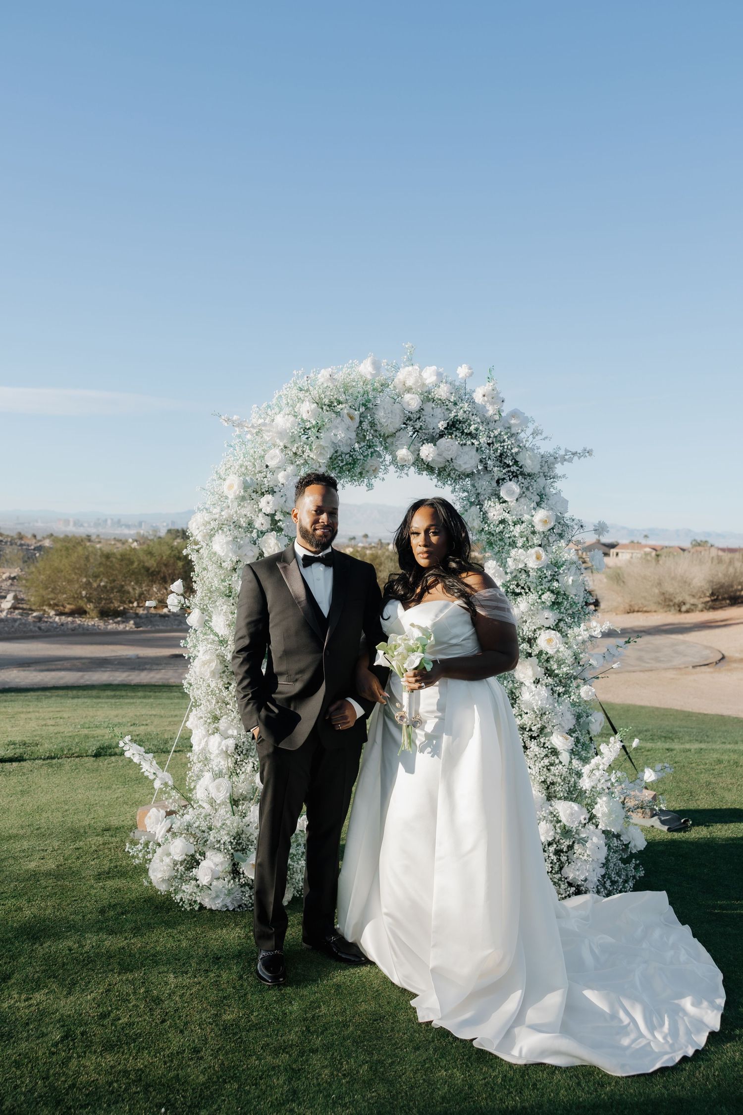 A bride and groom pose before a floral arch with white roses and greenery on a lush lawn under a clear blue sky.