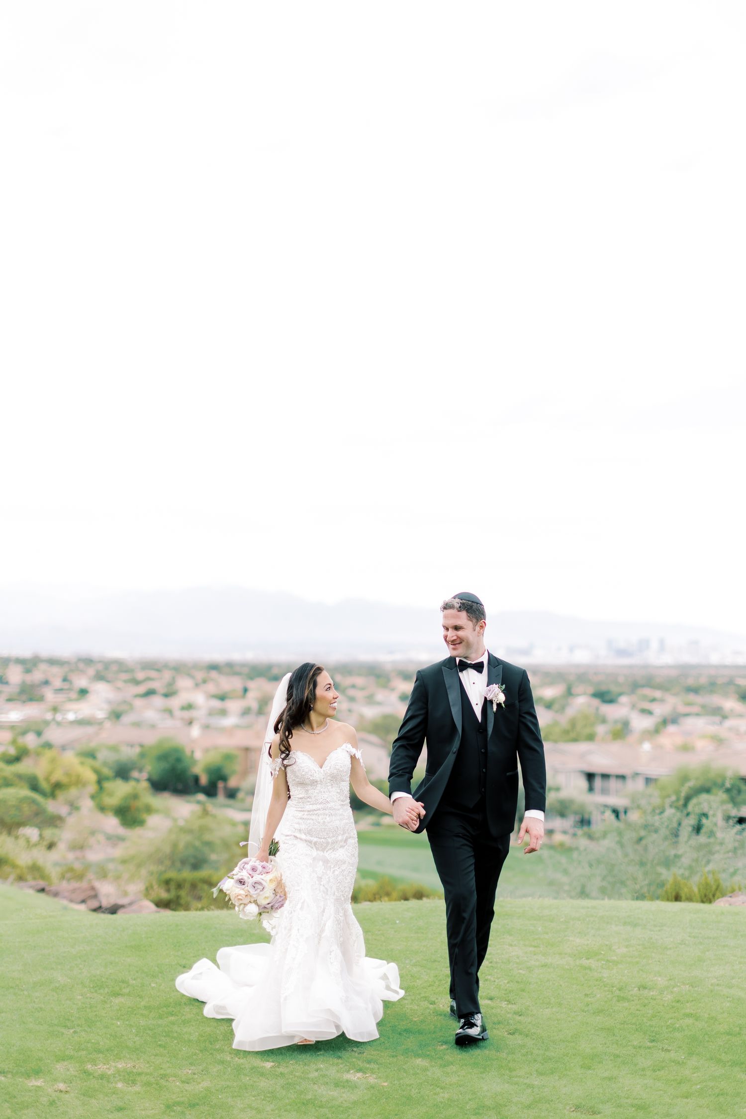 Bride and groom walk hand-in-hand on a golf course hilltop, with a scenic desert cityscape backdrop.