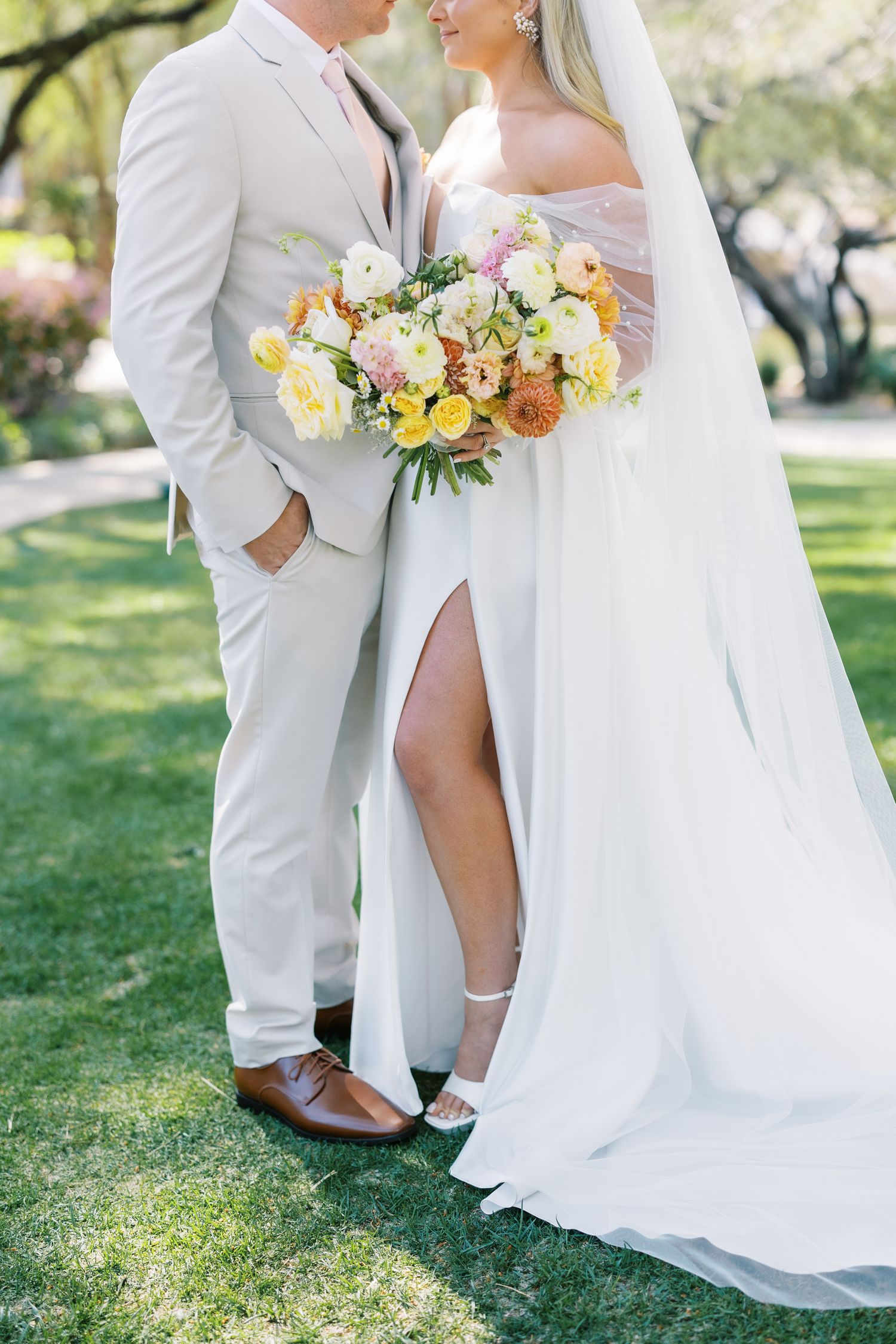 Bride and groom pose outdoors, she holds a yellow and pink bouquet in a white slit gown, he wears a tan suit.