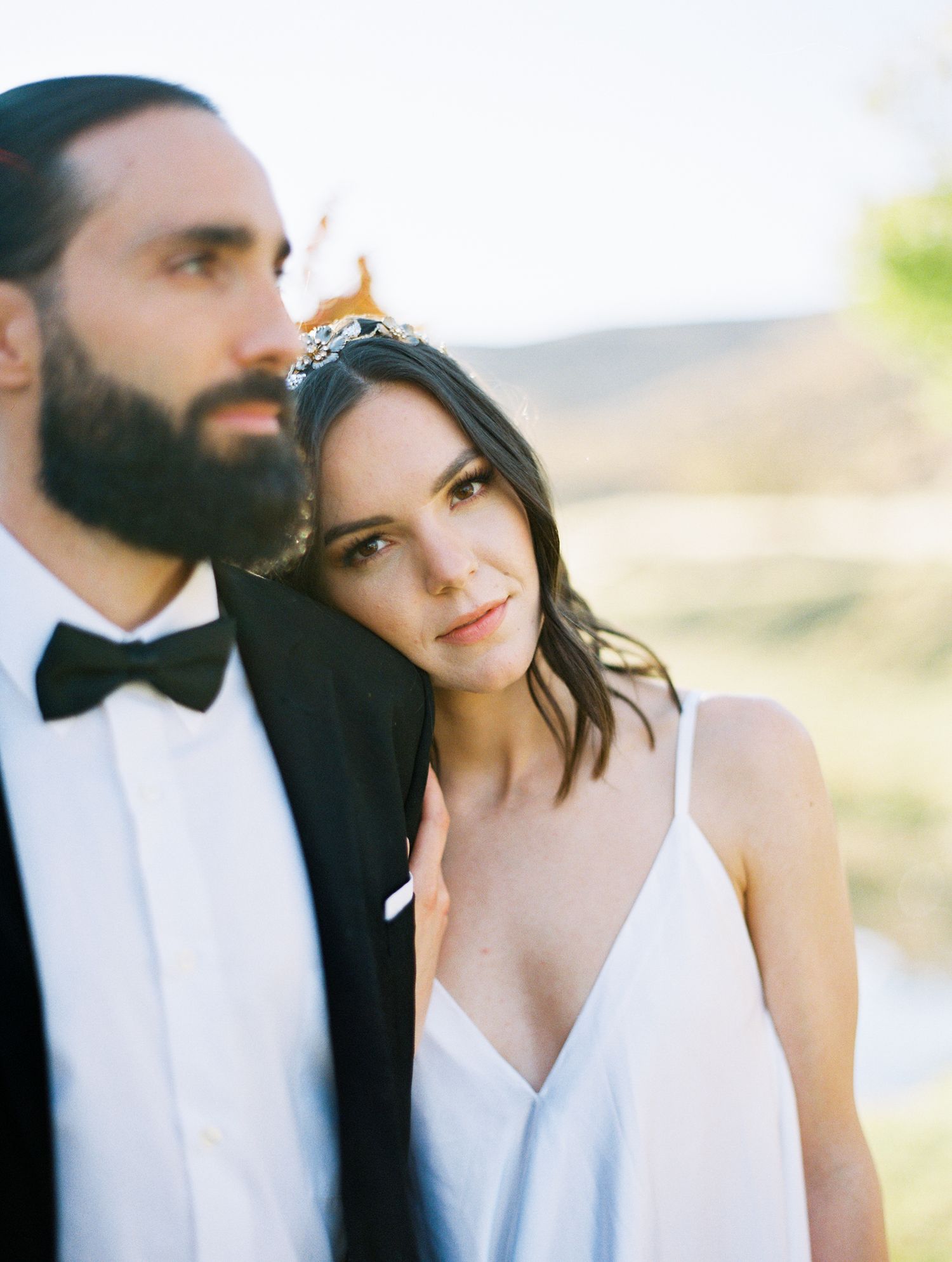 Elegant bride in white gown wearing tiara leans on groom in black tuxedo with bow tie outdoors.
