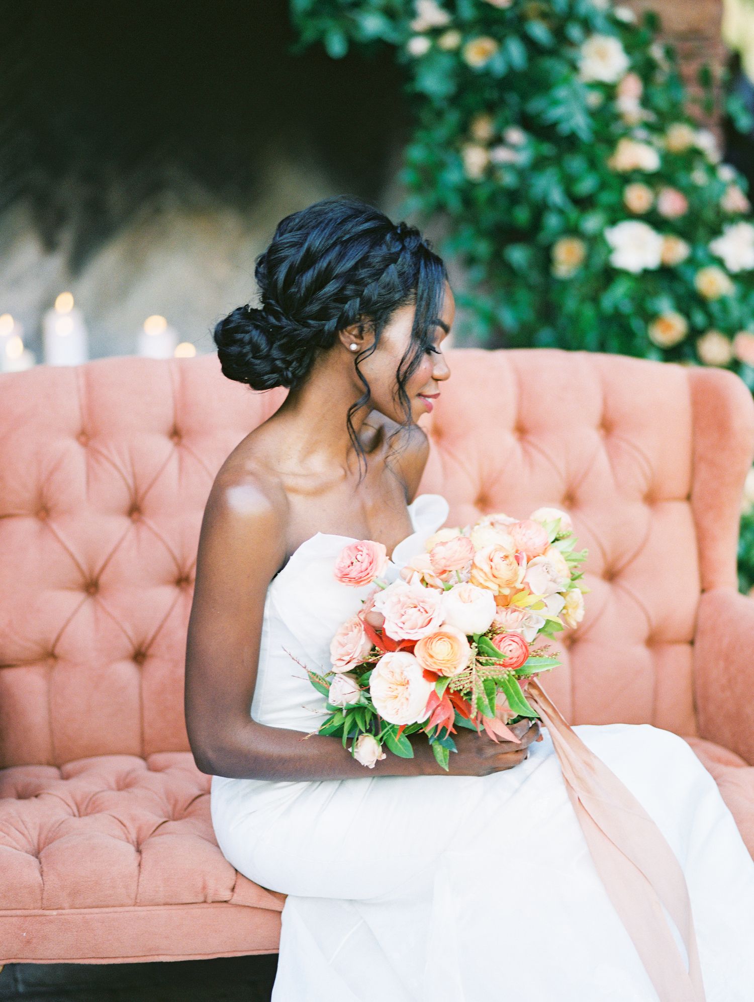Bride in white strapless gown sits on pink tufted sofa holding peach floral bouquet with braided updo hairstyle.