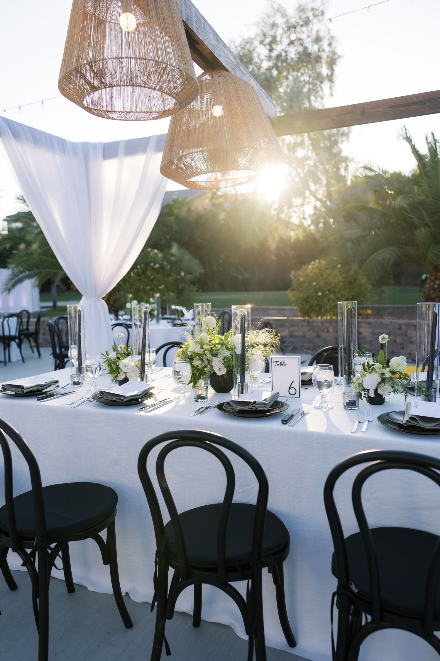 Elegant outdoor dining setup with white tablecloths, black chairs, and hanging wicker pendant lights at sunset.