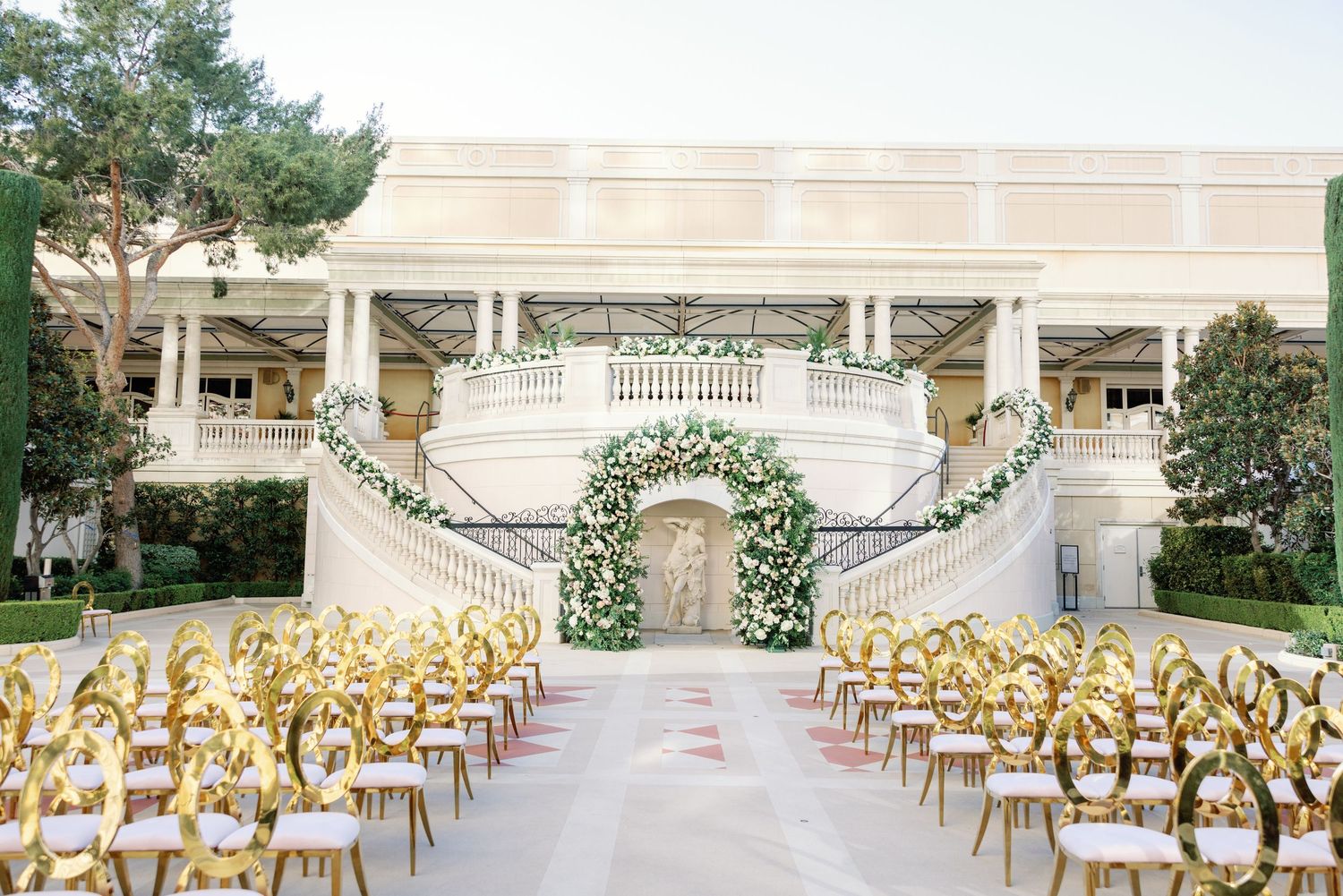Elegant outdoor wedding ceremony with gold chairs, floral arch, and grand white staircase venue.