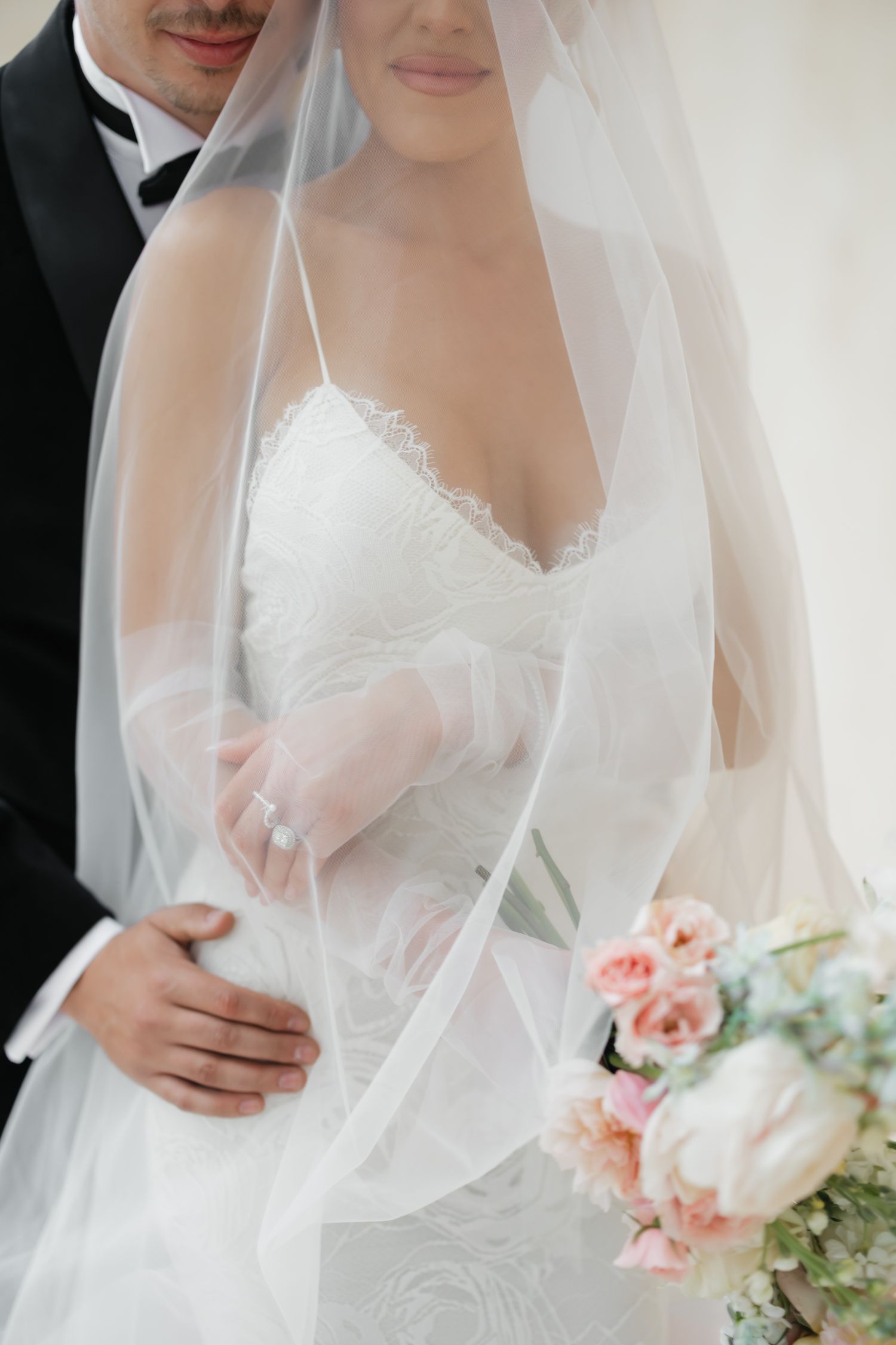 A bride in a white lace wedding dress holds a soft pink and green bouquet while wearing a flowing sheer veil.
