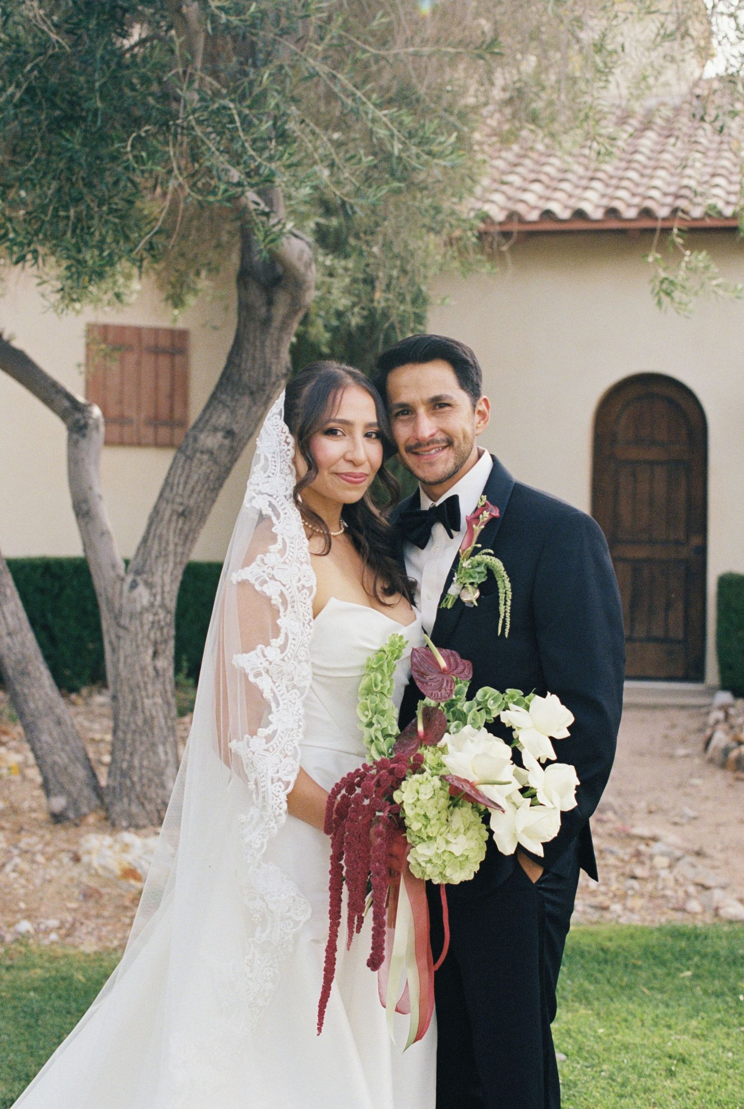 Bride and groom pose together outdoors, she holds a bouquet of white flowers and red amaranthus, wearing a veil and strapless gown.
