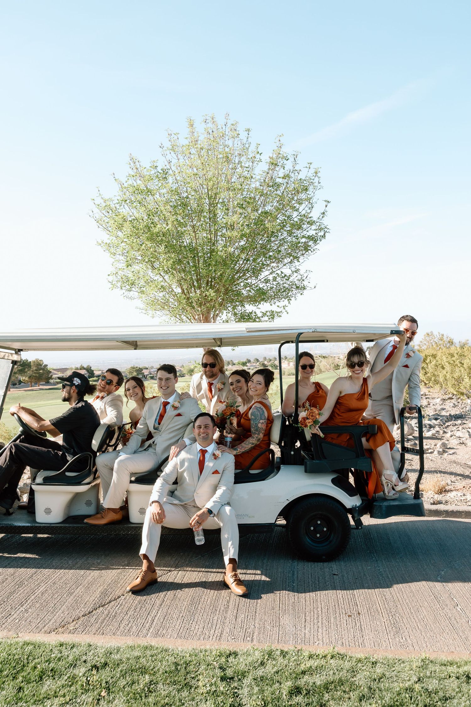 Wedding party posing on a large white golf cart outdoors, dressed in rust and cream formal attire on a sunny day.