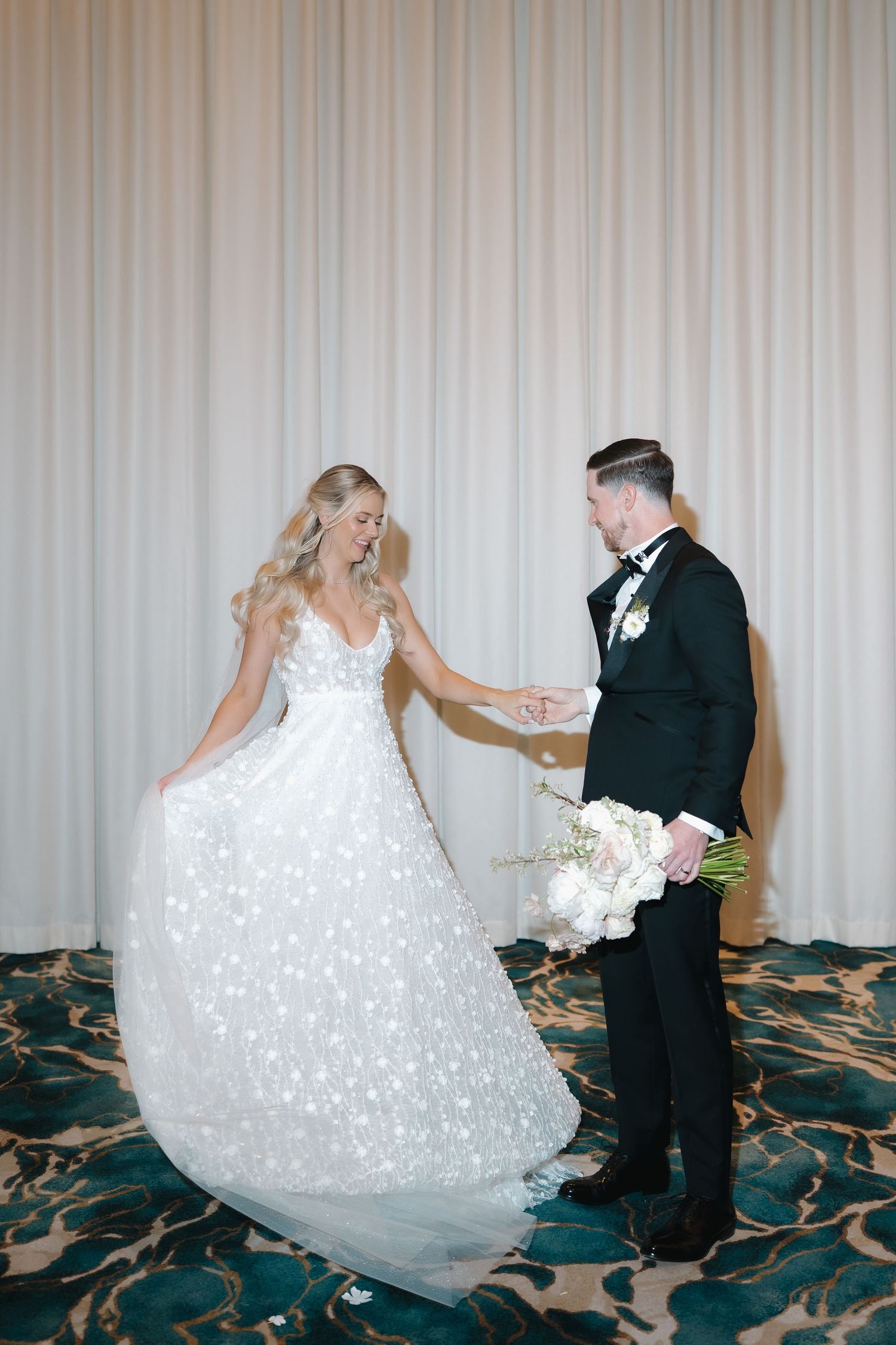 Bride in floral ballgown twirls while groom in black tuxedo holds her hand at elegant wedding reception.