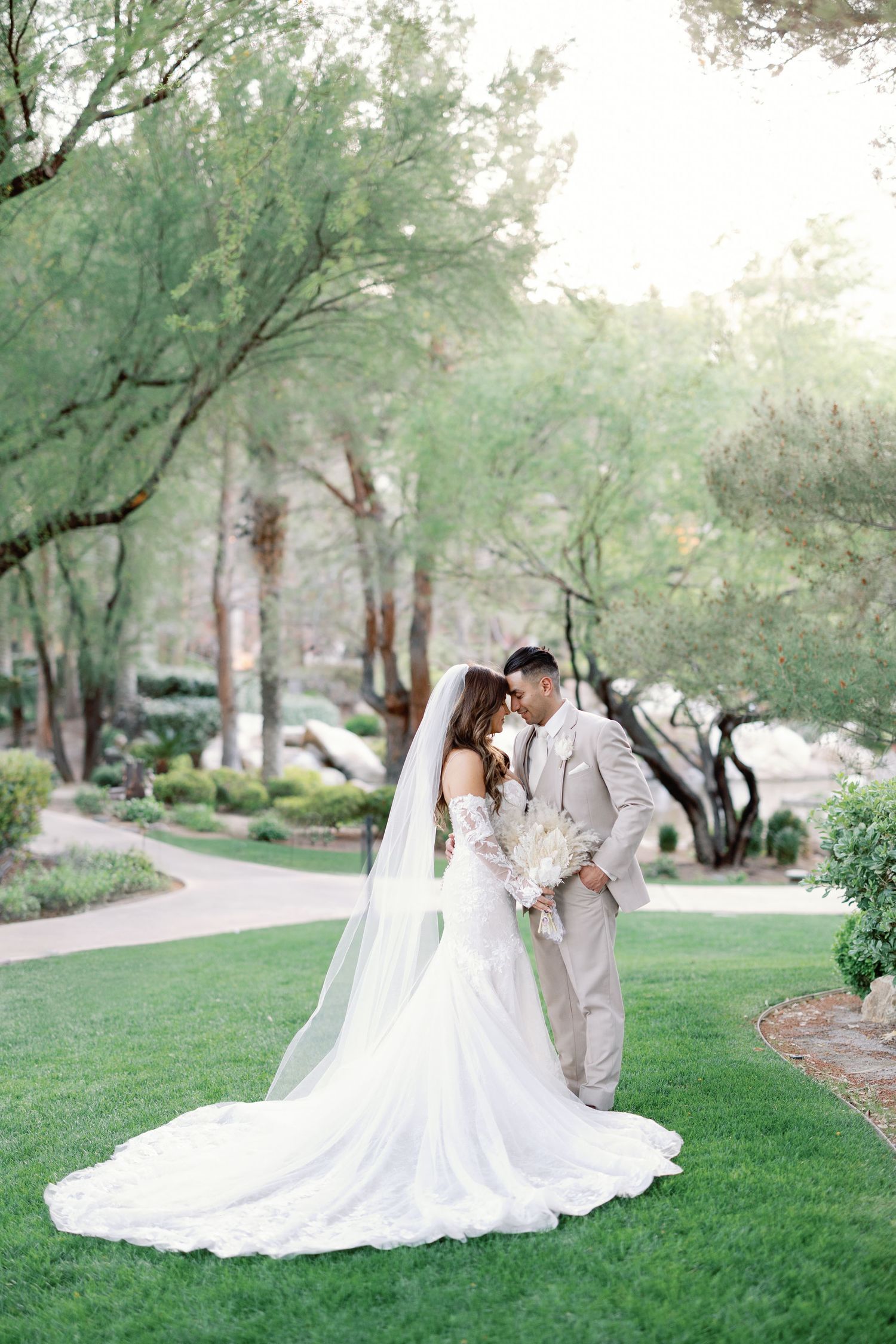 Bride and groom share a romantic moment on a green lawn surrounded by lush trees.