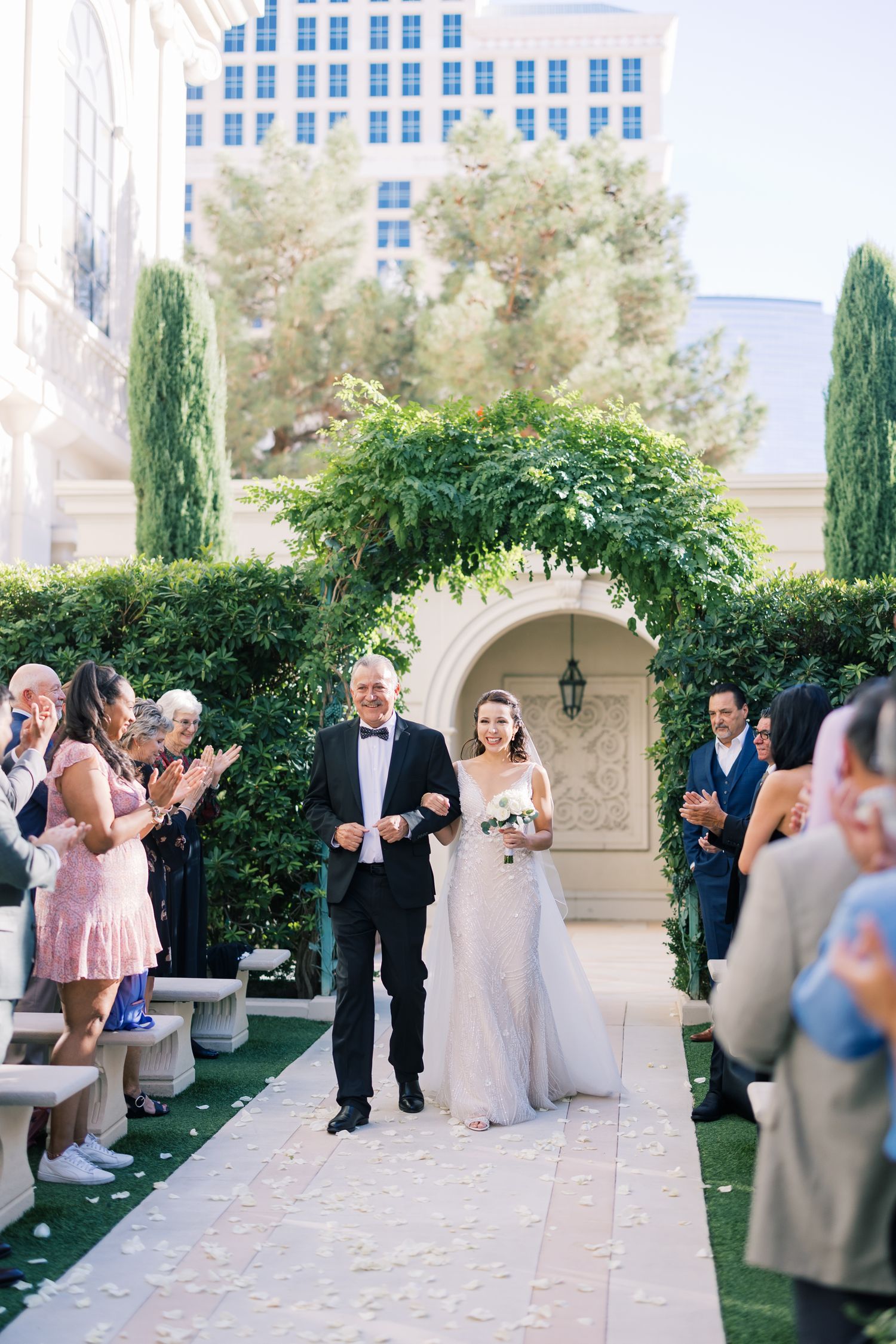 Bride walks down outdoor aisle with escort through floral arch, guests applauding at Las Vegas wedding.