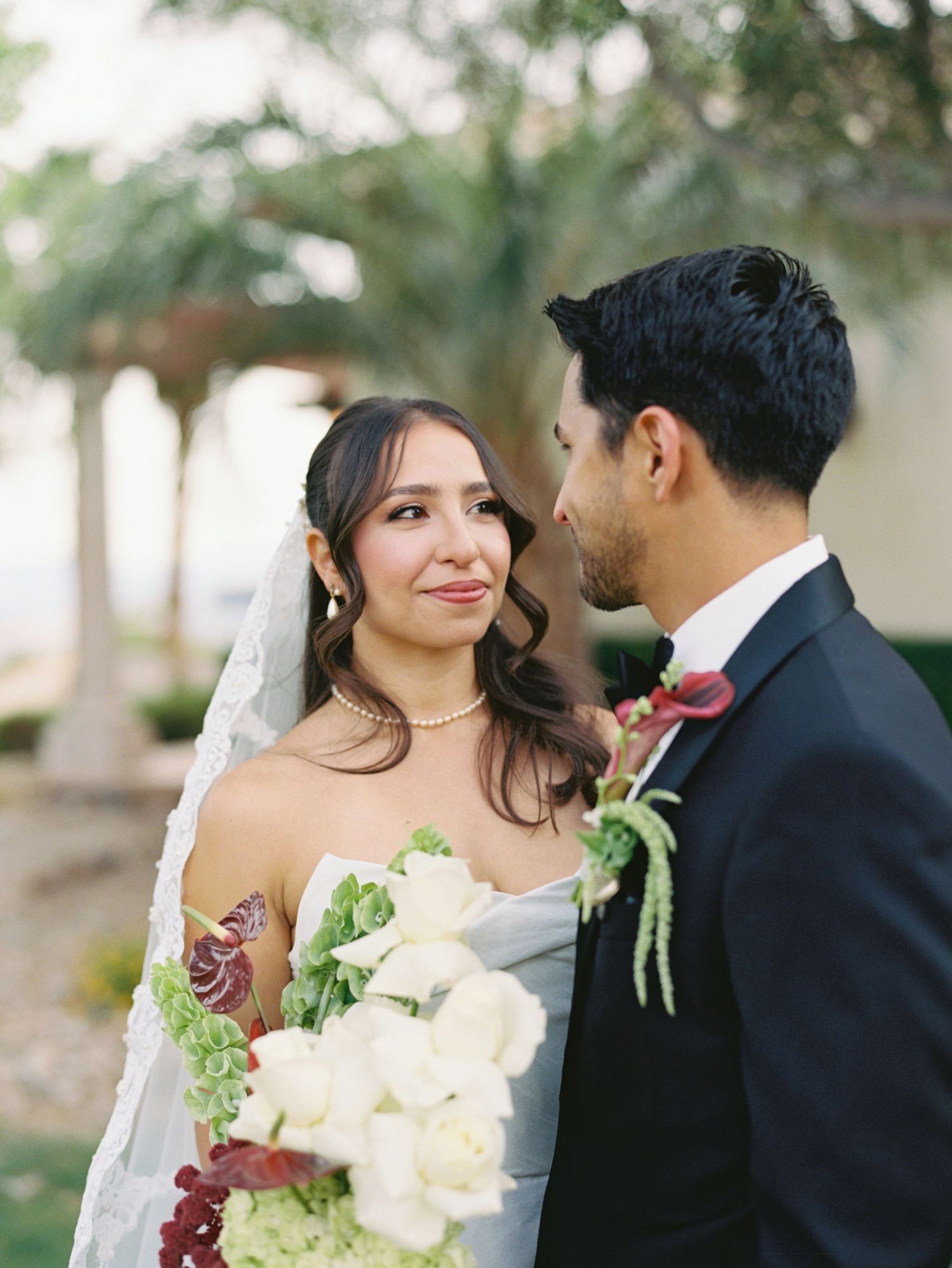 Bride holding white rose bouquet gazes at groom in dark suit with red boutonniere at outdoor wedding ceremony.