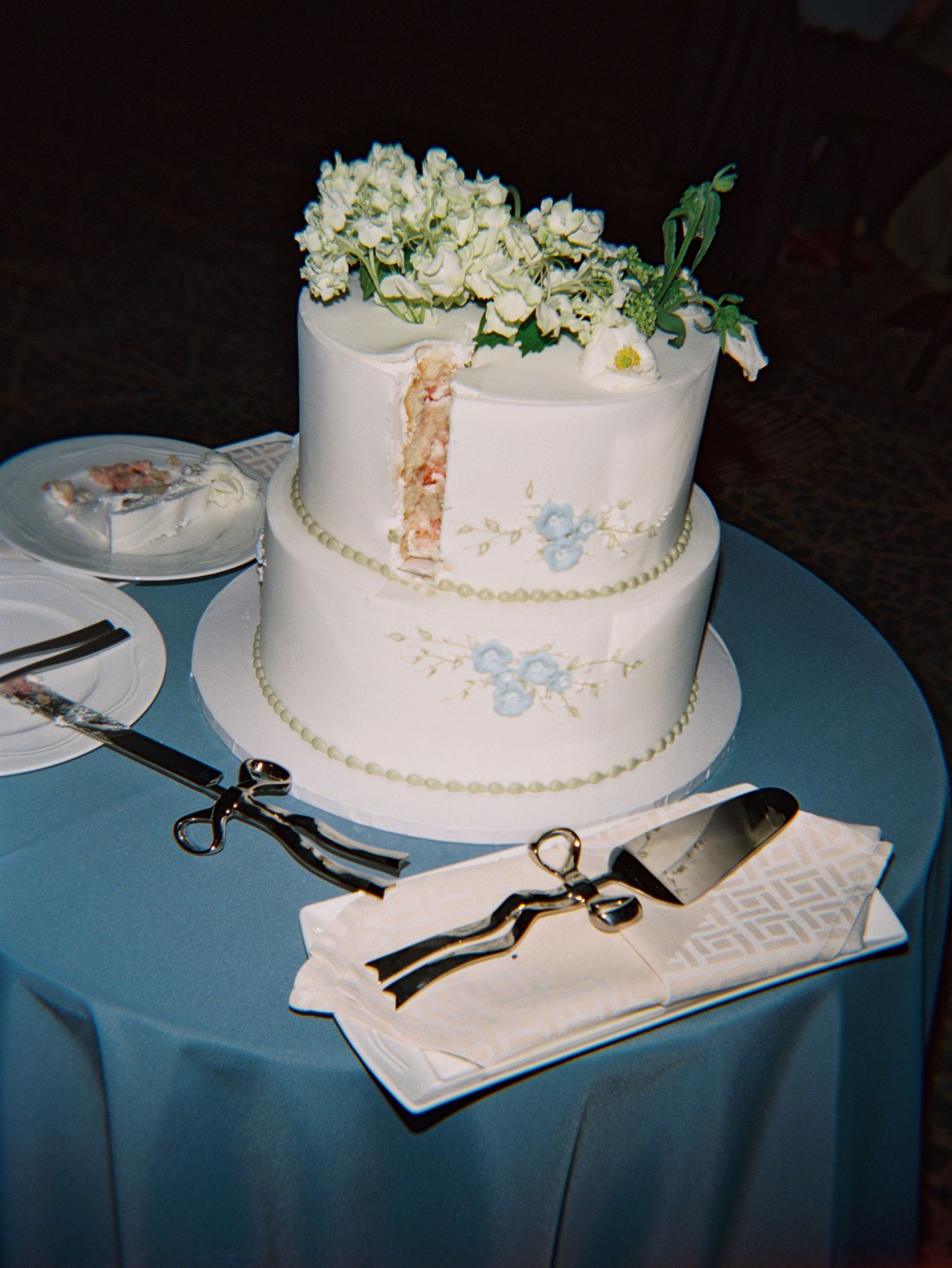Two-tier white wedding cake with blue floral details and fresh flowers on top, displayed with cake server and scissors on blue tablecloth.