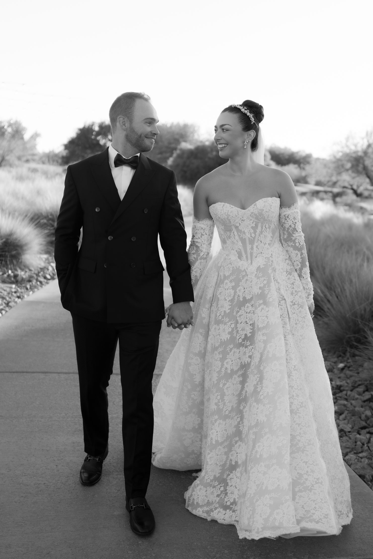 Black and white photo of a smiling bride in lace gown and groom in tuxedo holding hands walking outdoors.