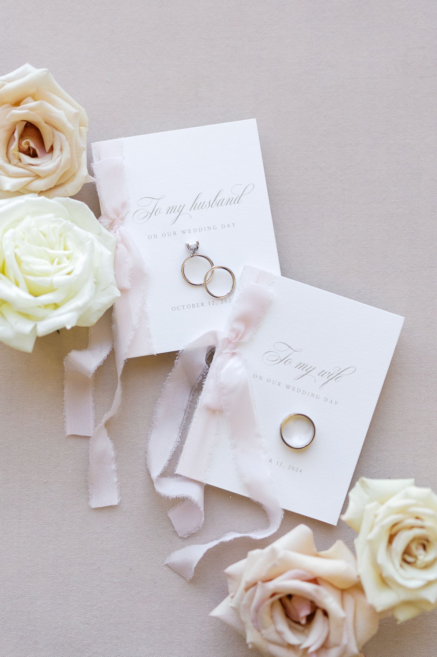 Elegant wedding flat lay with gold rings, vow cards, blush silk ribbon, and cream roses on a neutral background.