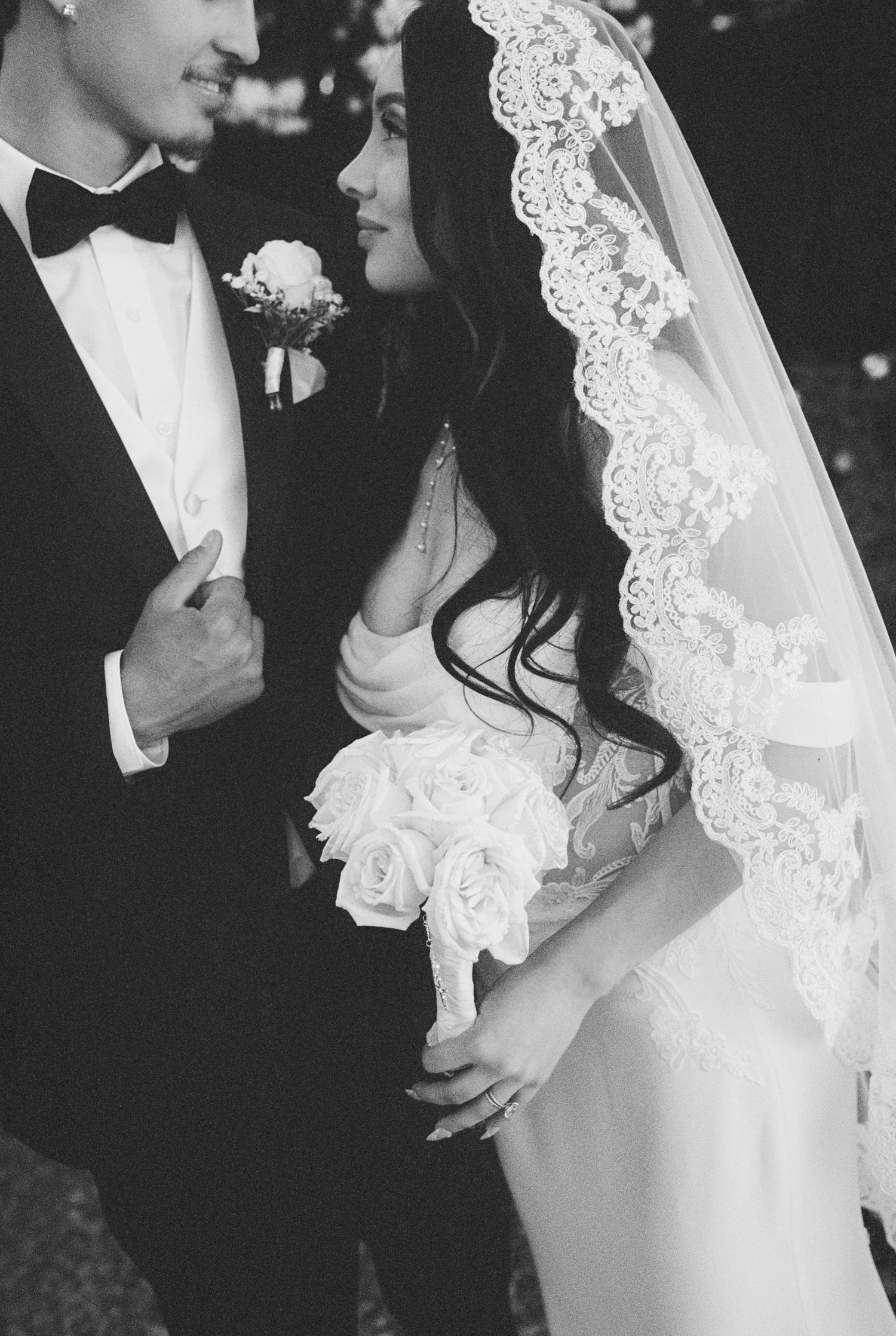 Black and white wedding photo of bride in lace veil holding roses bouquet standing close with groom in tuxedo.