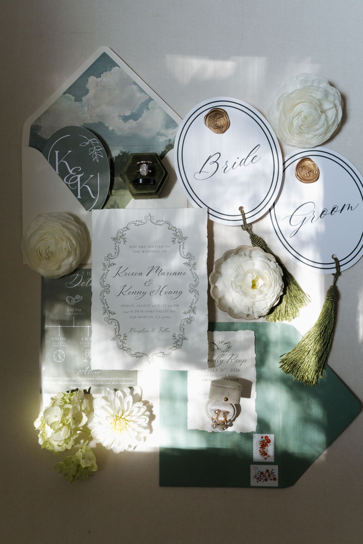 Elegant wedding stationery flat lay featuring green watercolor envelopes, floral invitations, bride and groom tags, and white flowers.