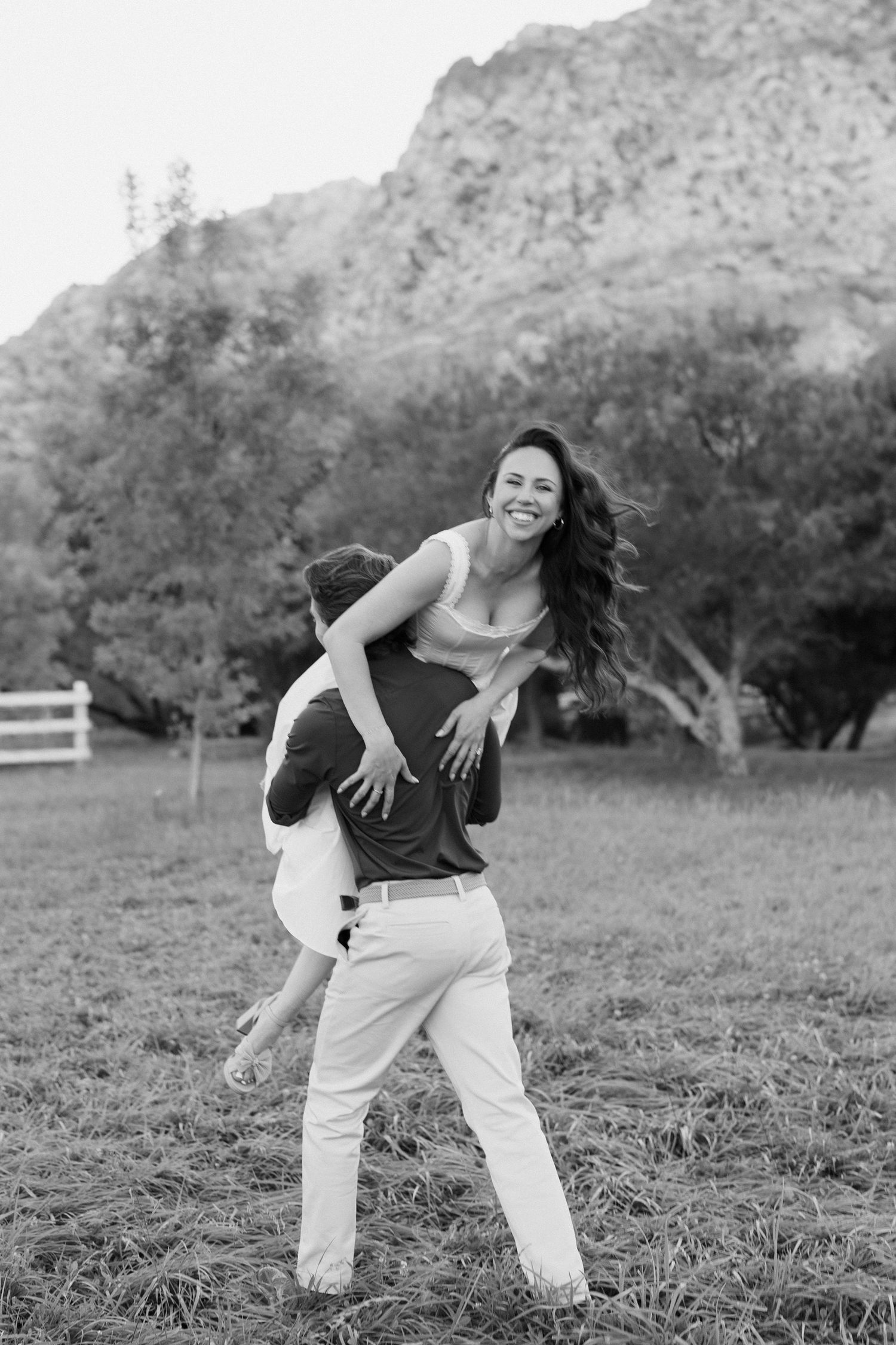 Black and white photo of a man lifting a laughing woman outdoors with mountains and trees in the background.