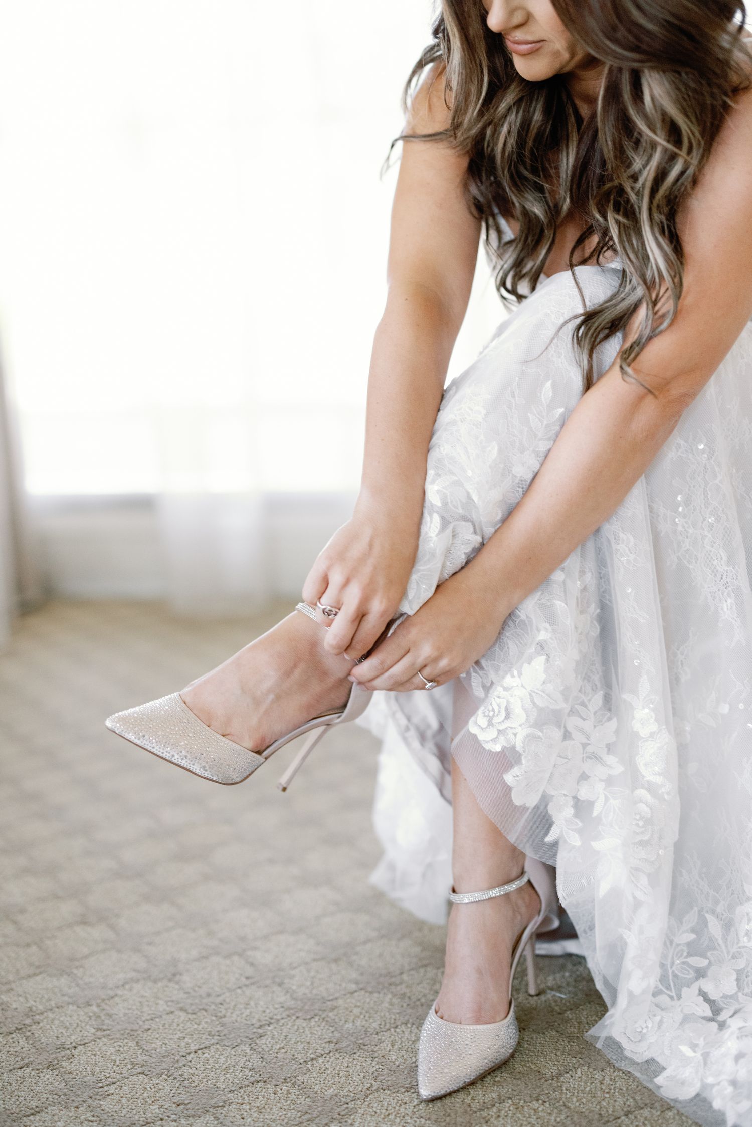 Bride in lace wedding dress putting on sparkly ivory heeled shoes with ankle strap before ceremony.