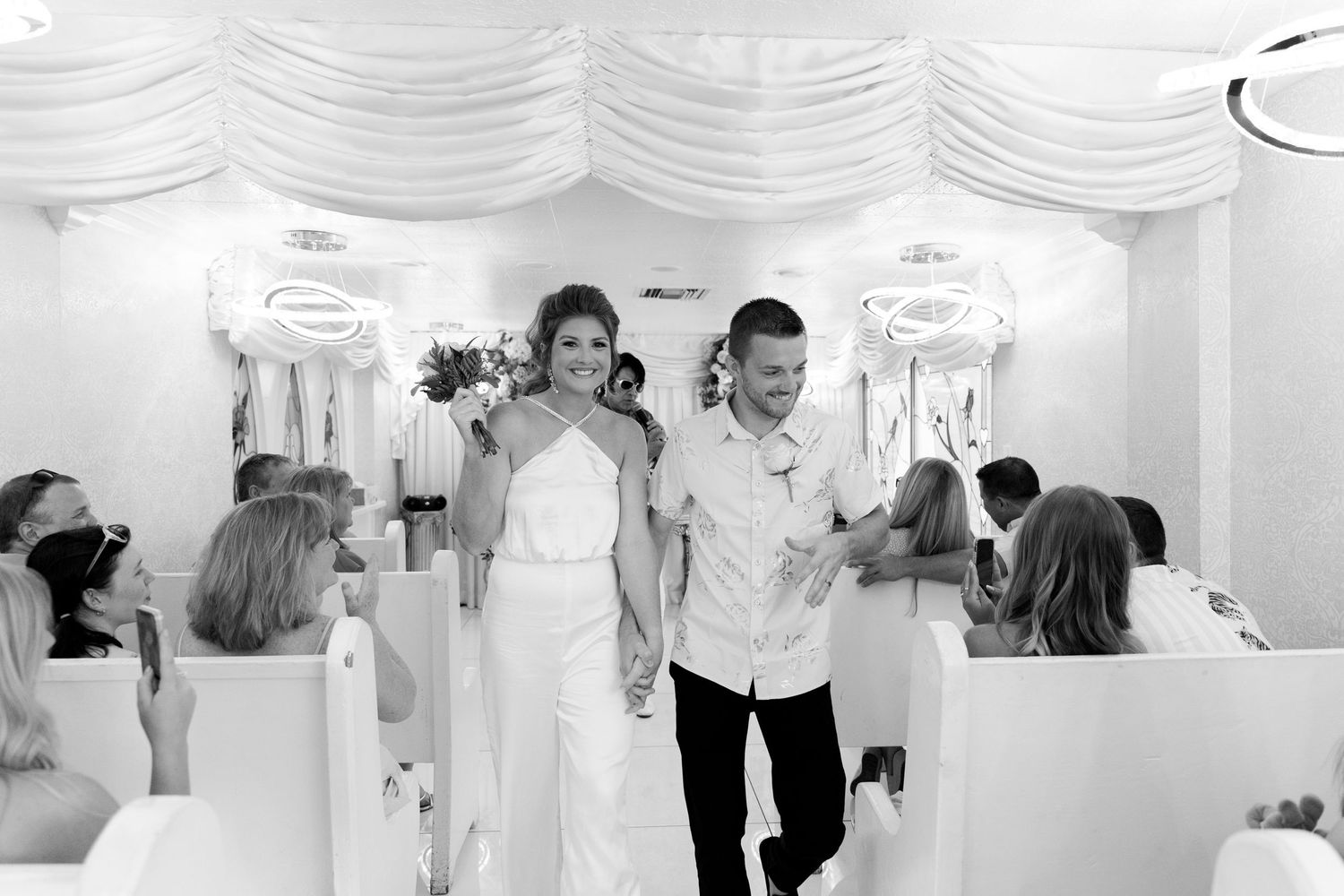 Black and white photo of a smiling bride and groom walking down the aisle after their wedding ceremony.