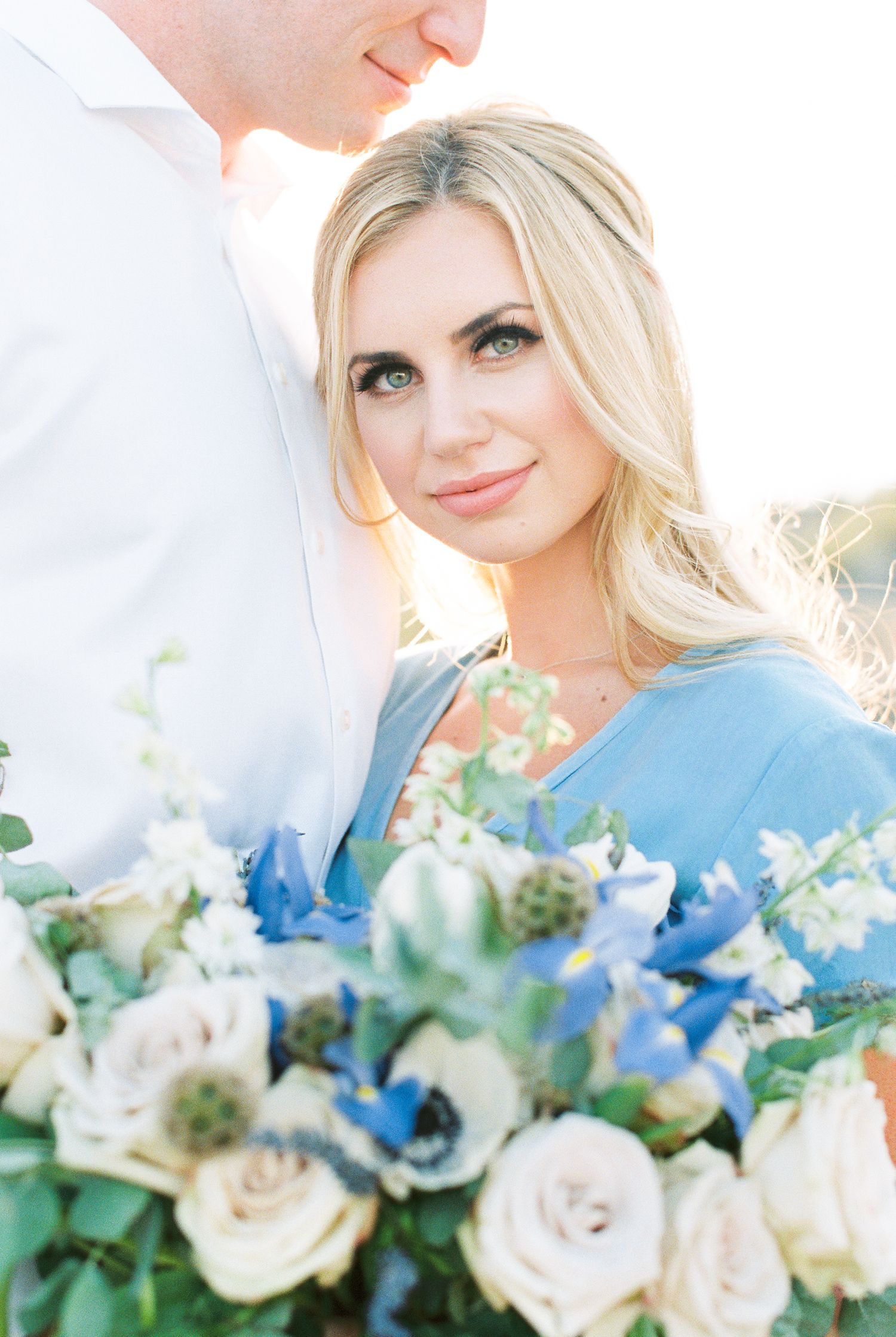 Blonde woman in blue dress holds white and blue floral bouquet during romantic outdoor engagement photo session.