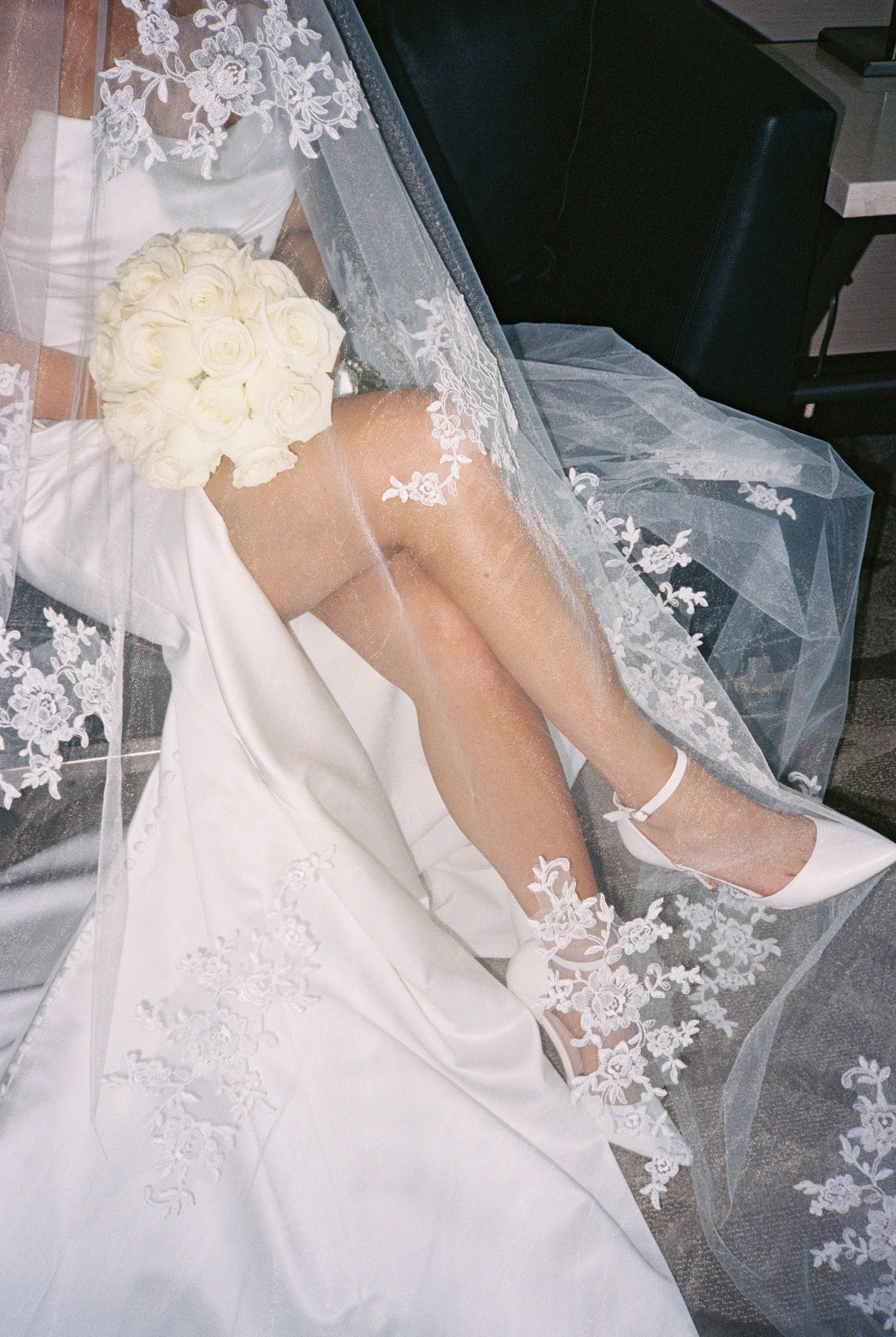 Bride in white lace wedding dress and veil, holding white rose bouquet, wearing white heels with ankle strap.