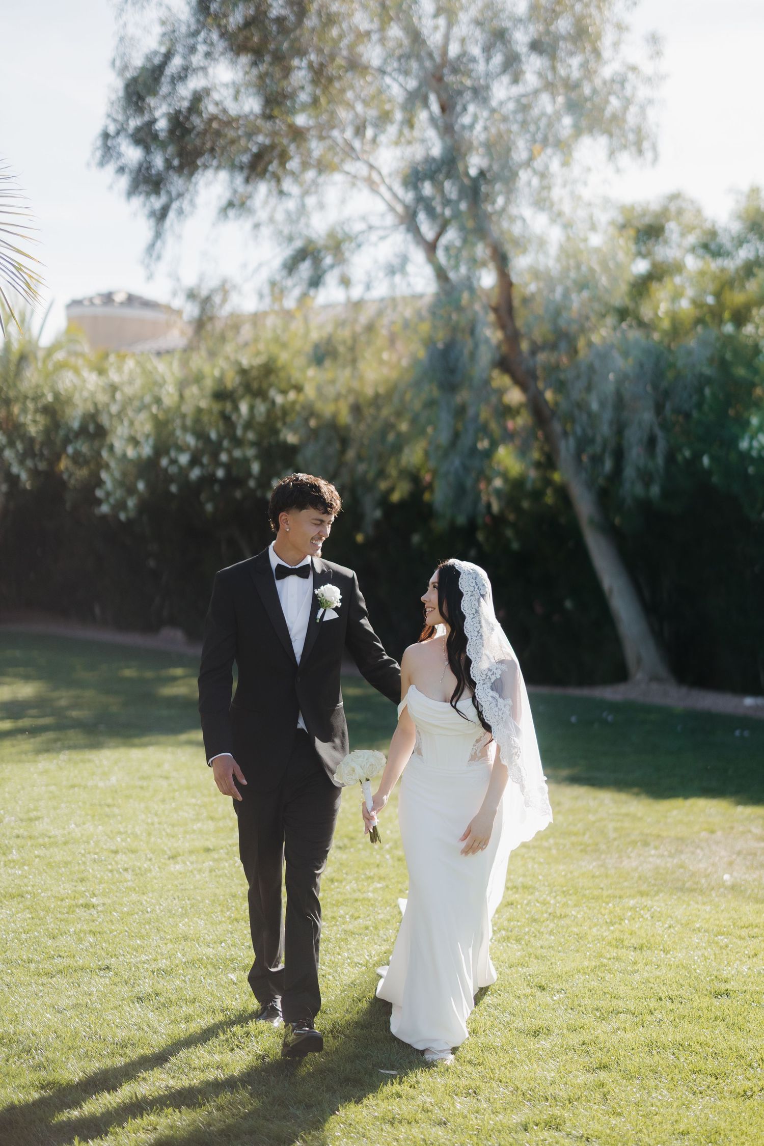 Bride and groom walk hand in hand on green lawn, smiling at each other in elegant wedding attire.
