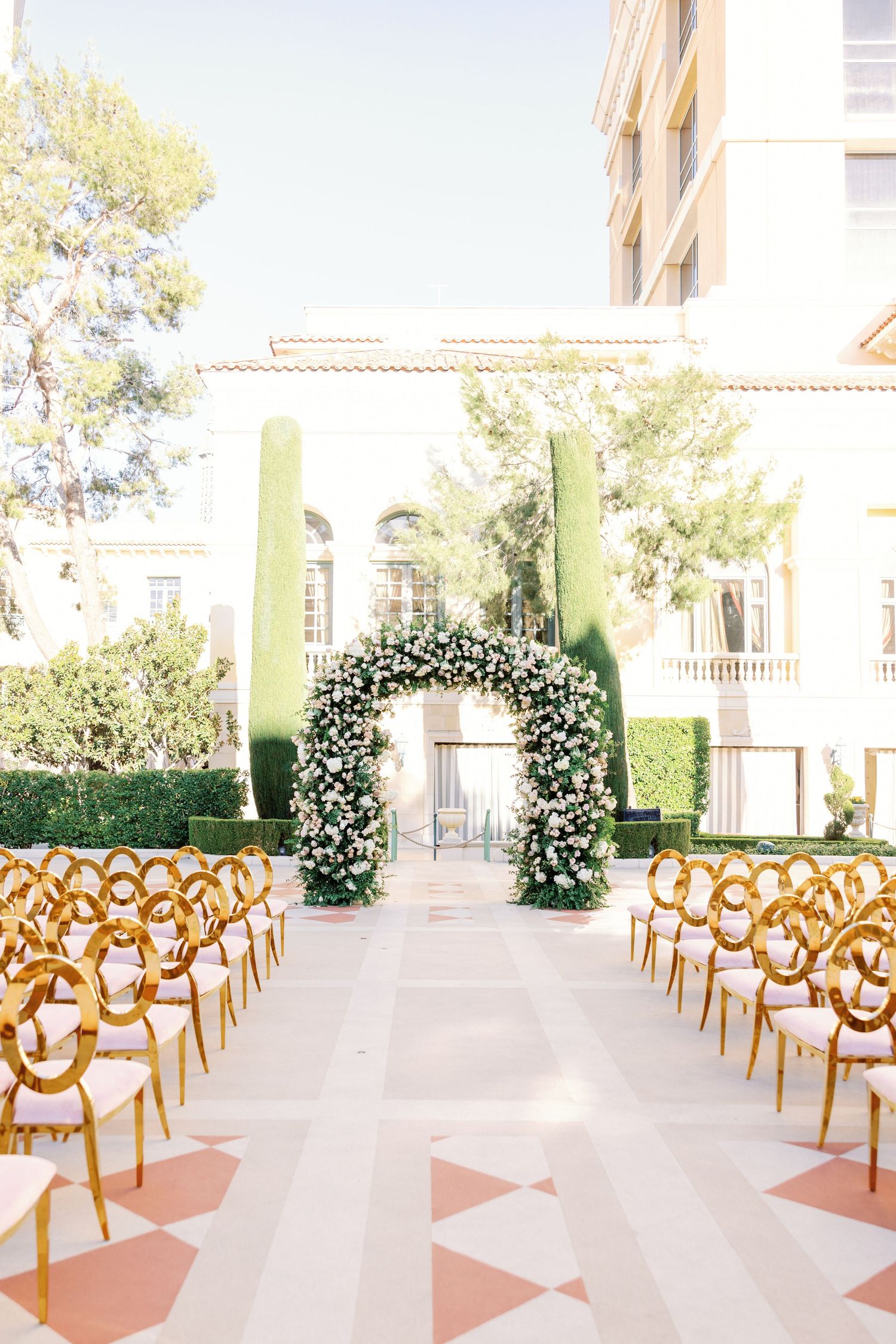 Outdoor wedding ceremony with gold chairs flanking a lush green and white floral arch at a luxury estate venue.