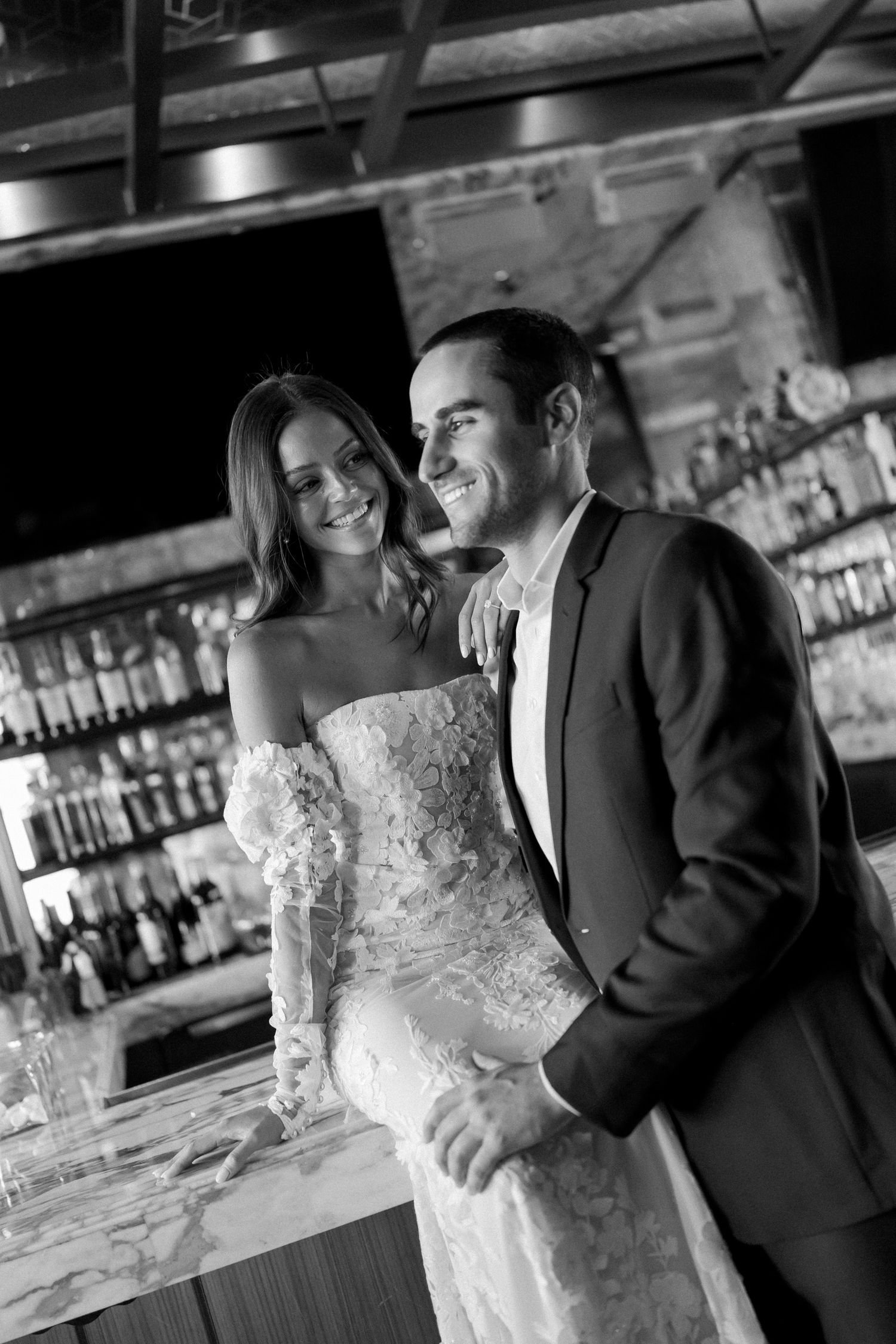 Black and white photo of a smiling bride and groom posing together at a bar on their wedding day.