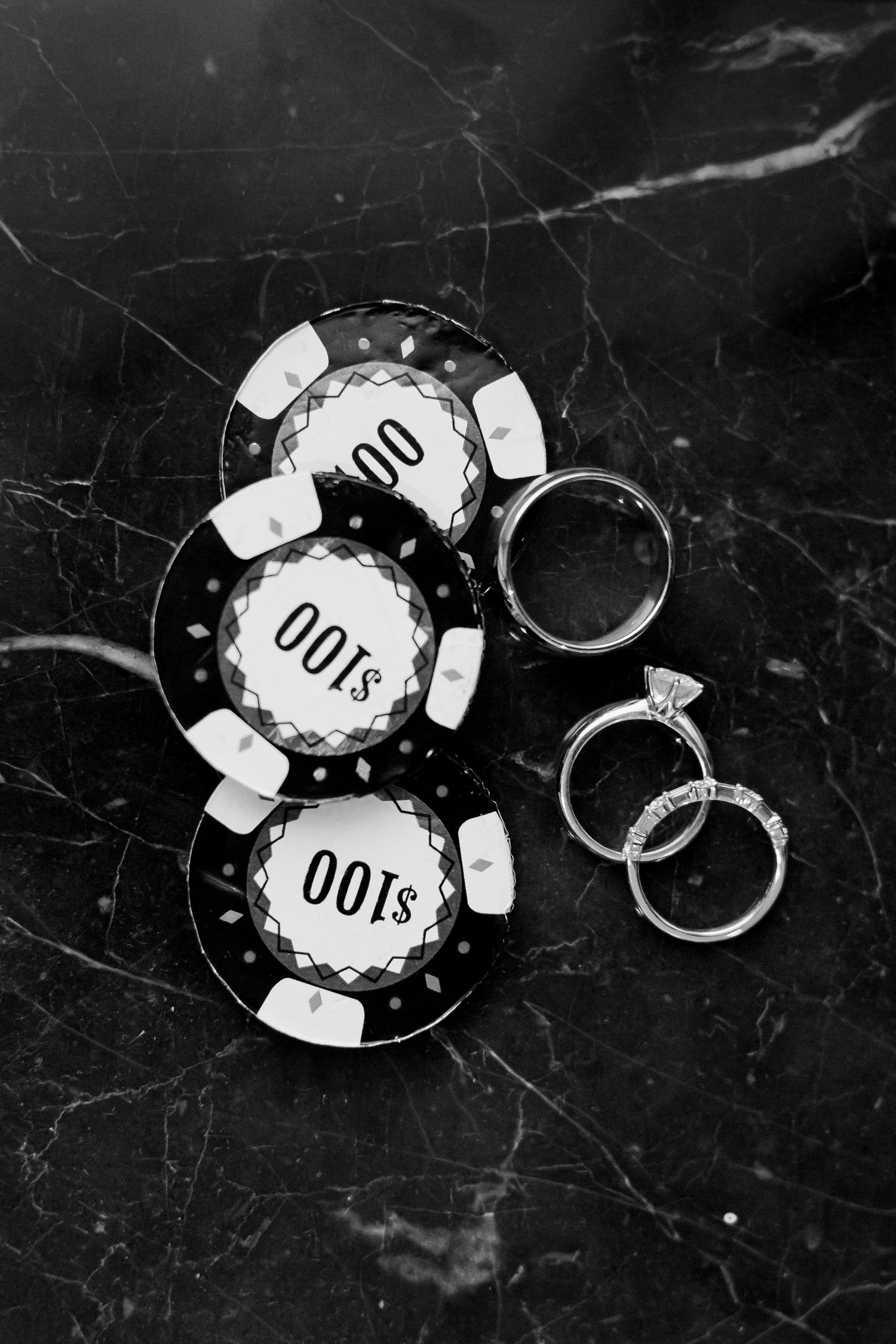 Black and white photo of $100 poker chips beside diamond engagement ring and wedding bands on marble surface.