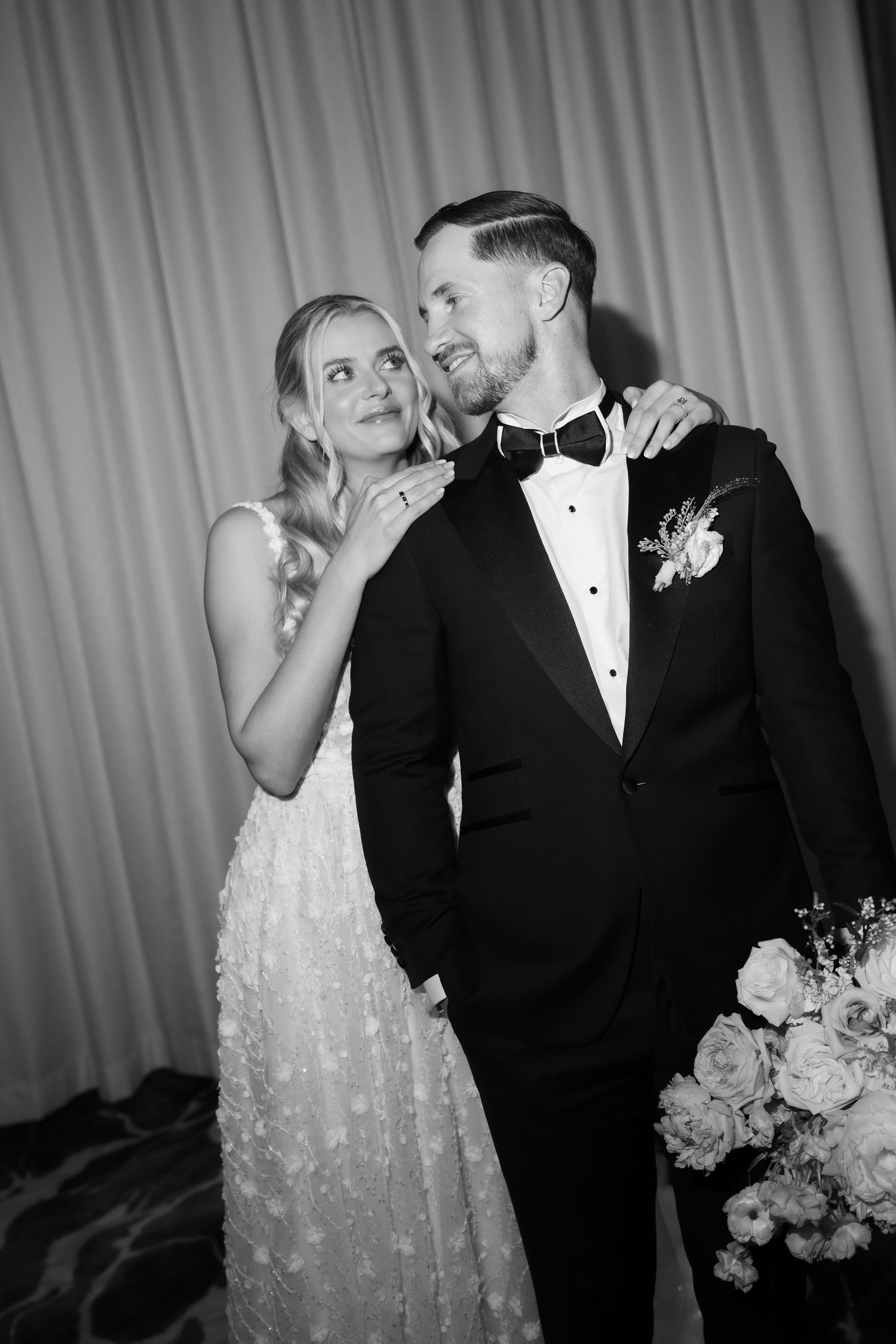 Black and white wedding photo of a bride in lace gown and groom in tuxedo with bow tie posing together.