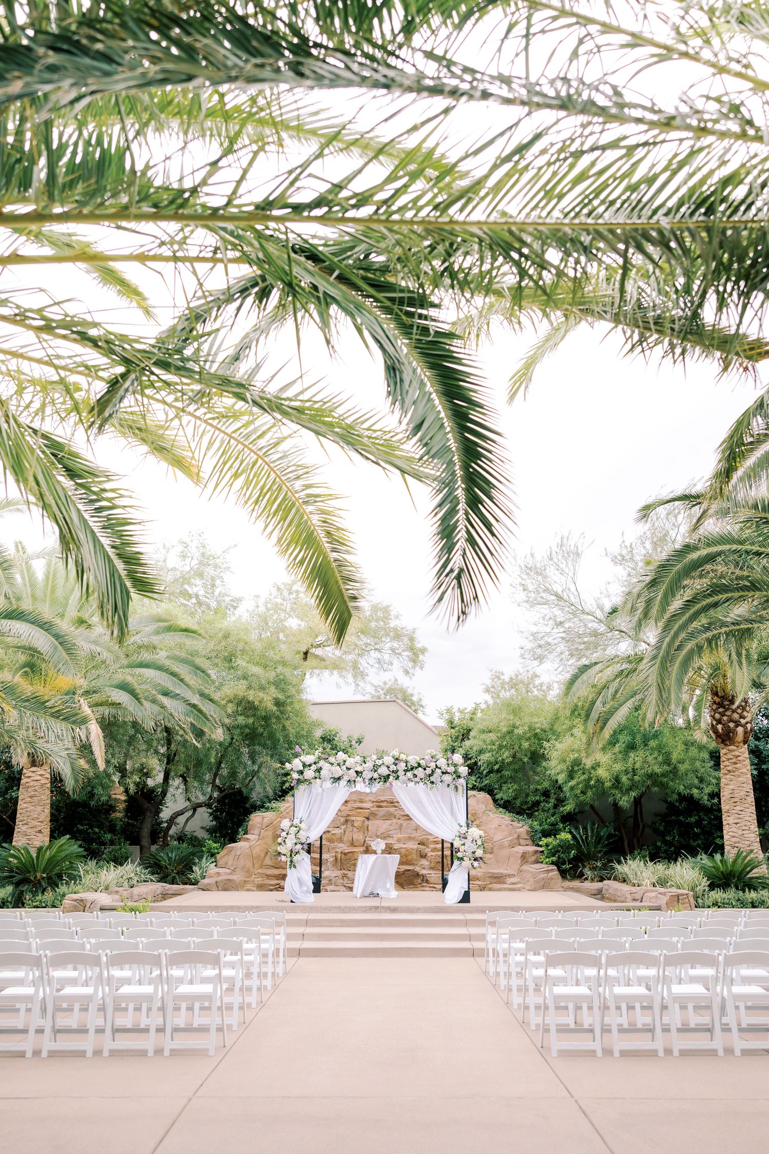 Outdoor wedding ceremony setup with white chairs, floral arch, and lush palm trees in a garden setting.