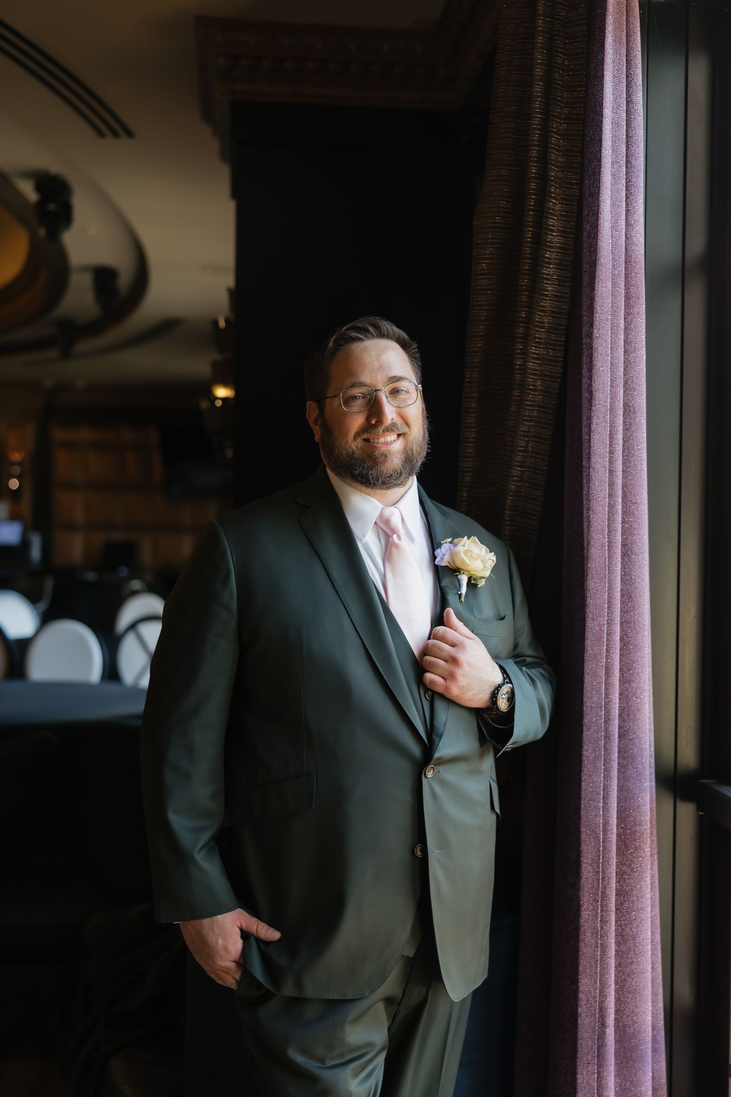 Smiling groom in green suit with boutonniere leaning against purple curtain in elegant venue.