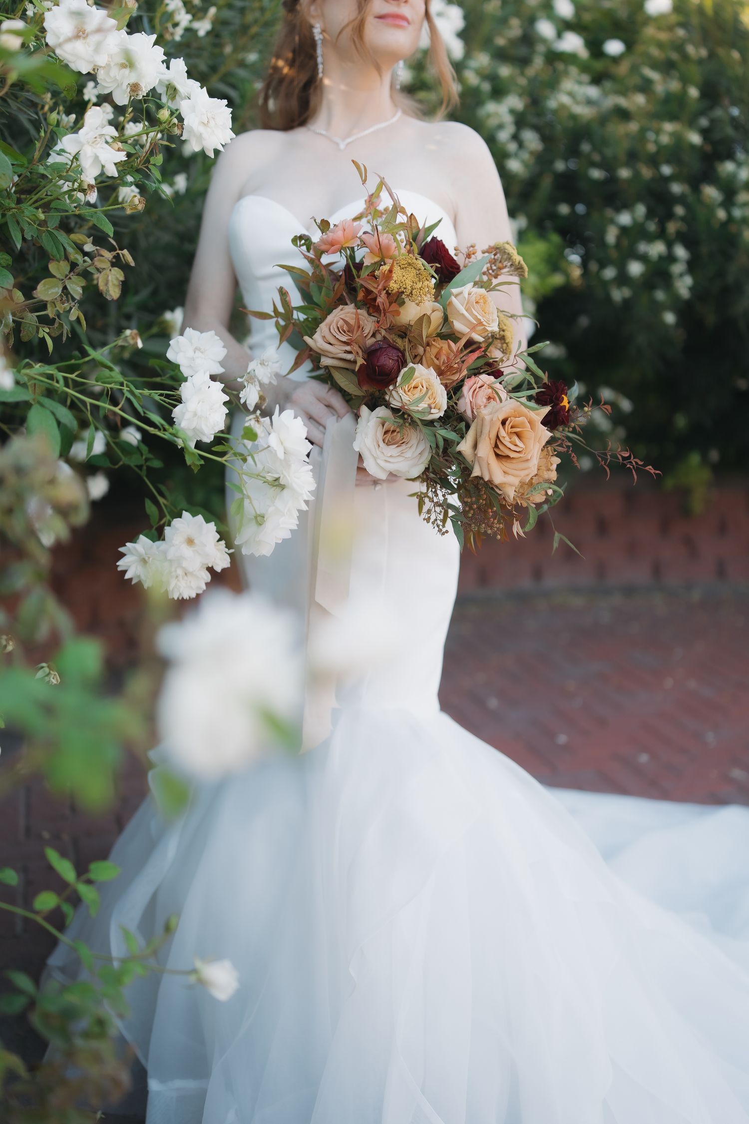 Bride in strapless mermaid wedding gown holding lush bouquet of roses in warm autumn tones outdoors.