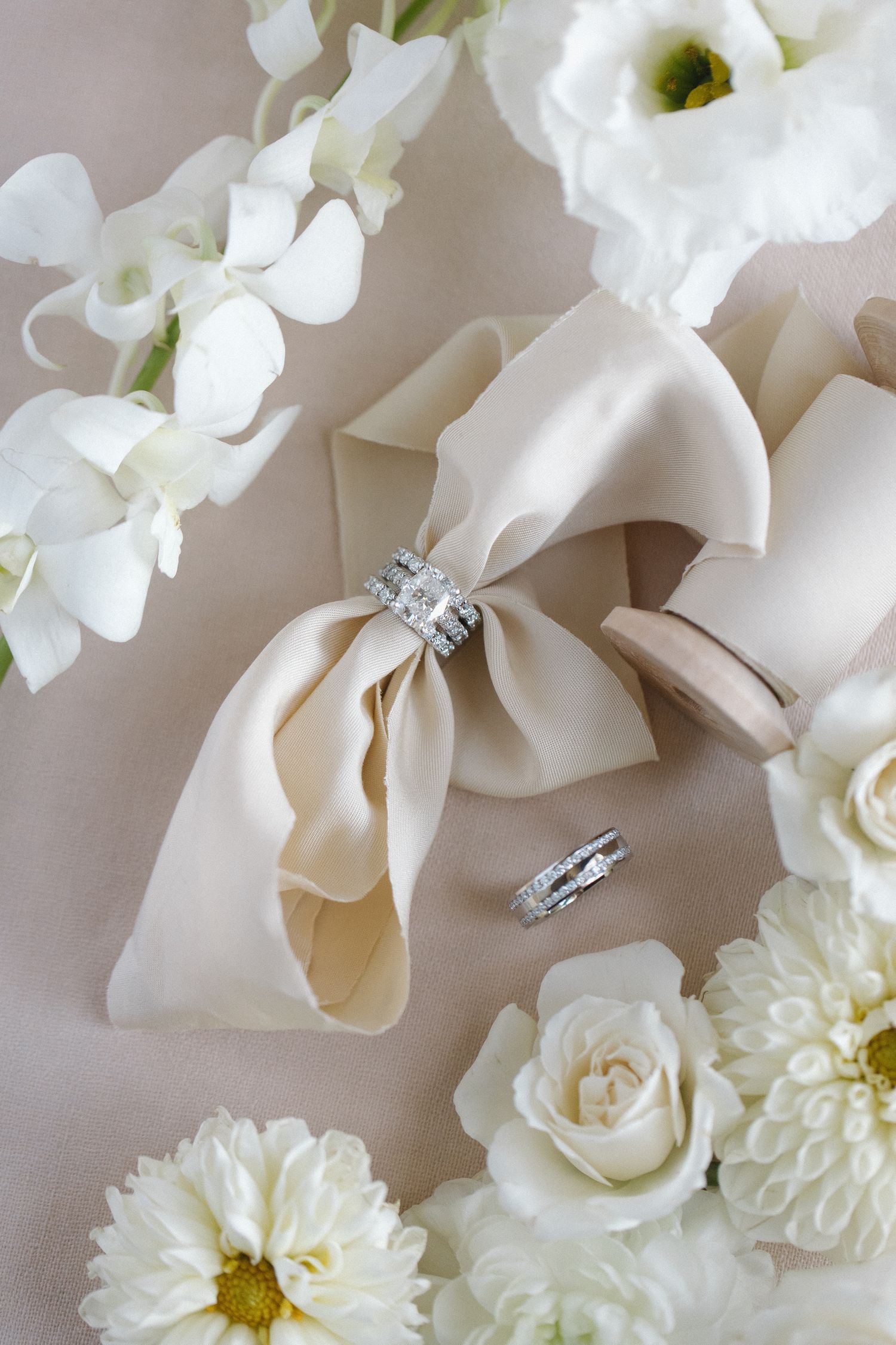 Wedding details featuring a cream silk bow, diamond rings, and white roses and flowers arranged on a light beige background.