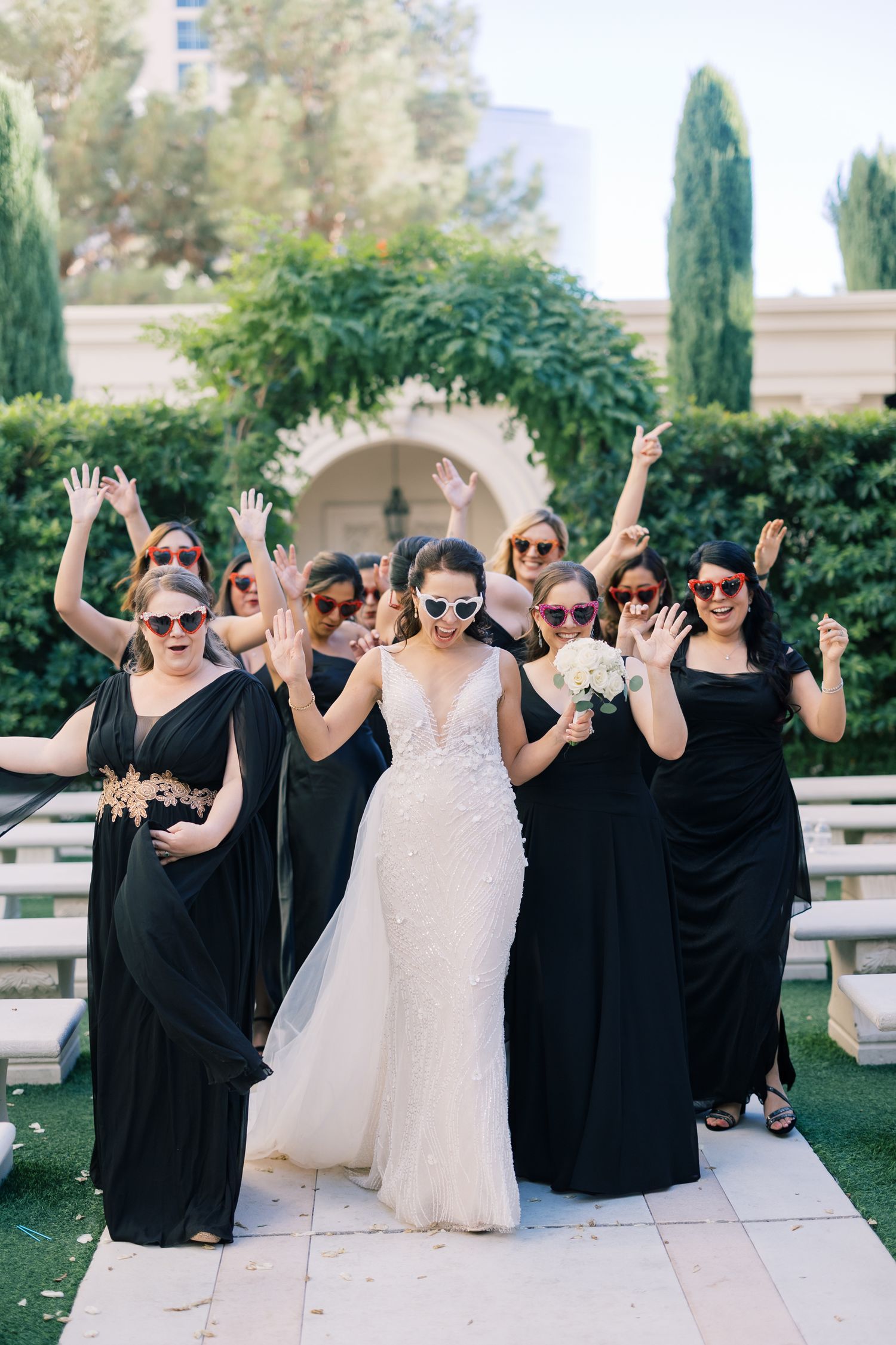Bride in lace gown celebrates with bridesmaids in black dresses wearing fun sunglasses in outdoor garden setting.