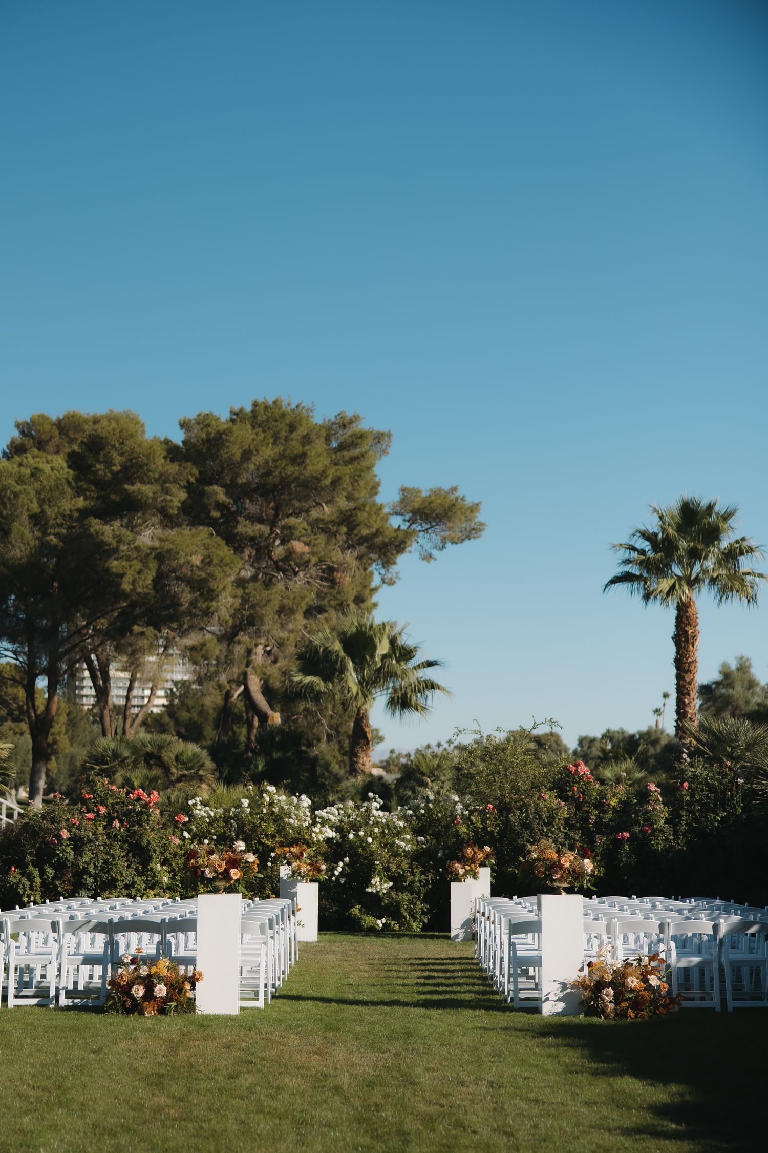 Elegant outdoor wedding ceremony setup with white chairs lining grass aisle, floral arrangements, and palm trees.