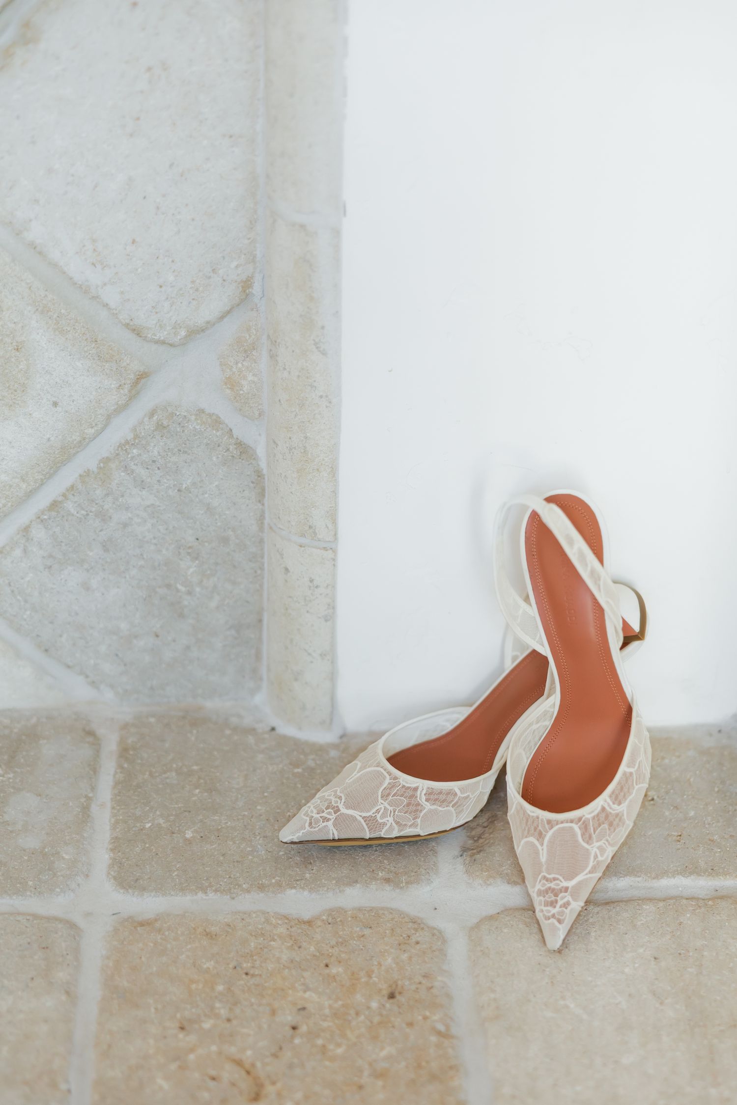 Elegant white lace slingback heels with pointed toes and tan insoles resting on stone floor near white wall.